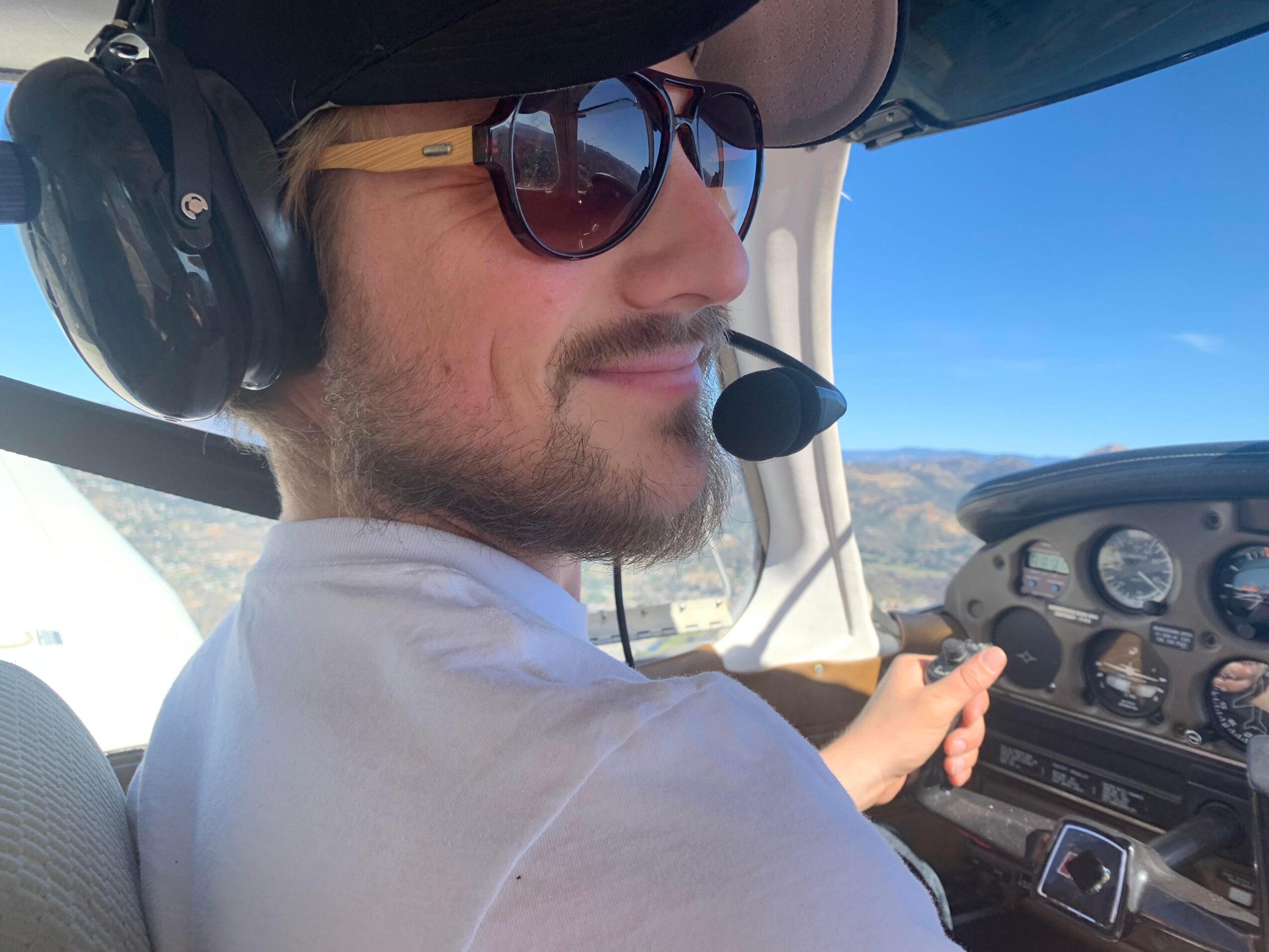 Man wearing sunglasses and headset, sitting in cockpit, smiling, with landscape and blue sky outside window.