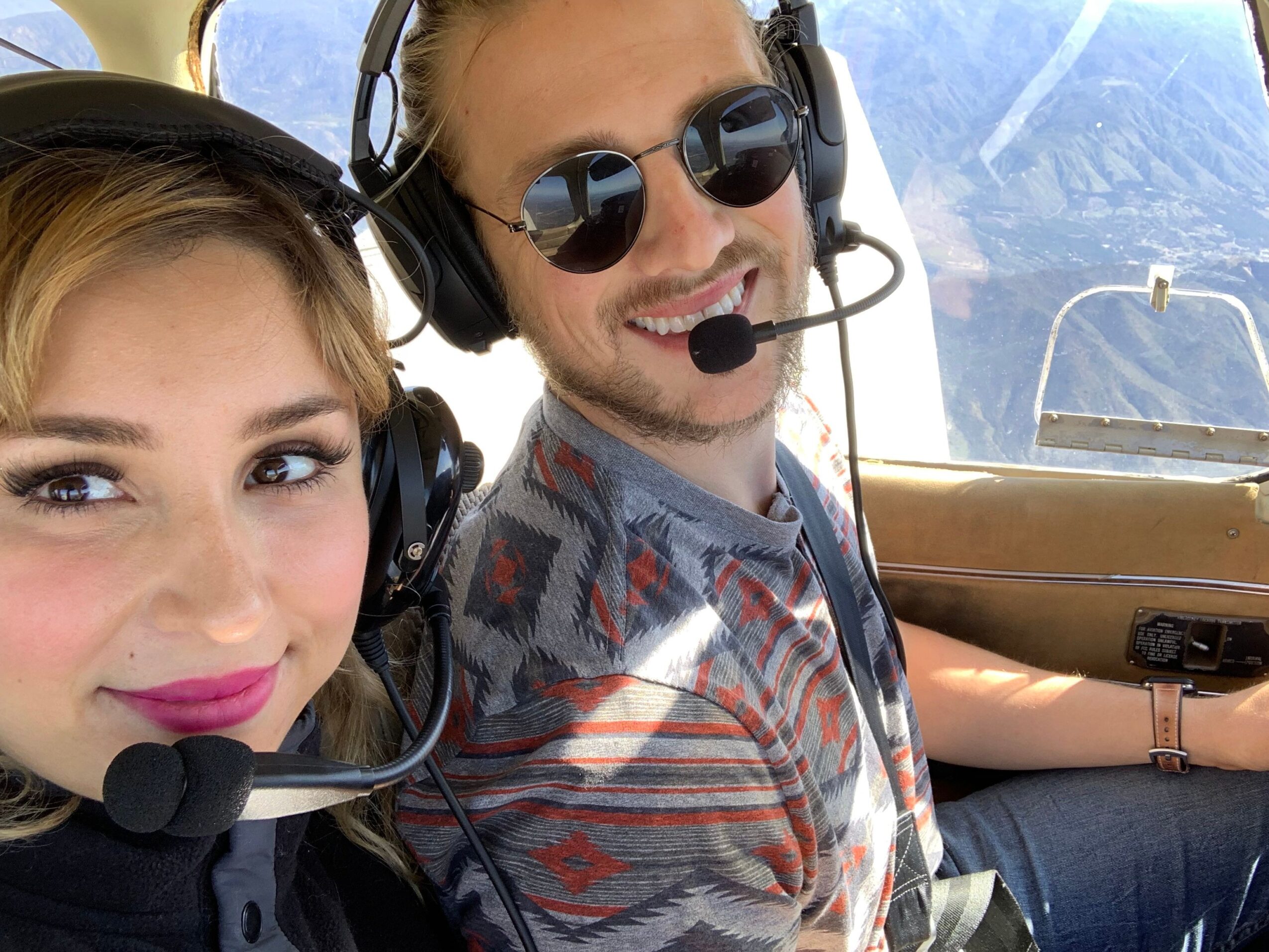 Two women wearing headsets, one with sunglasses, smiling, with a blue sky and clouds in the background.