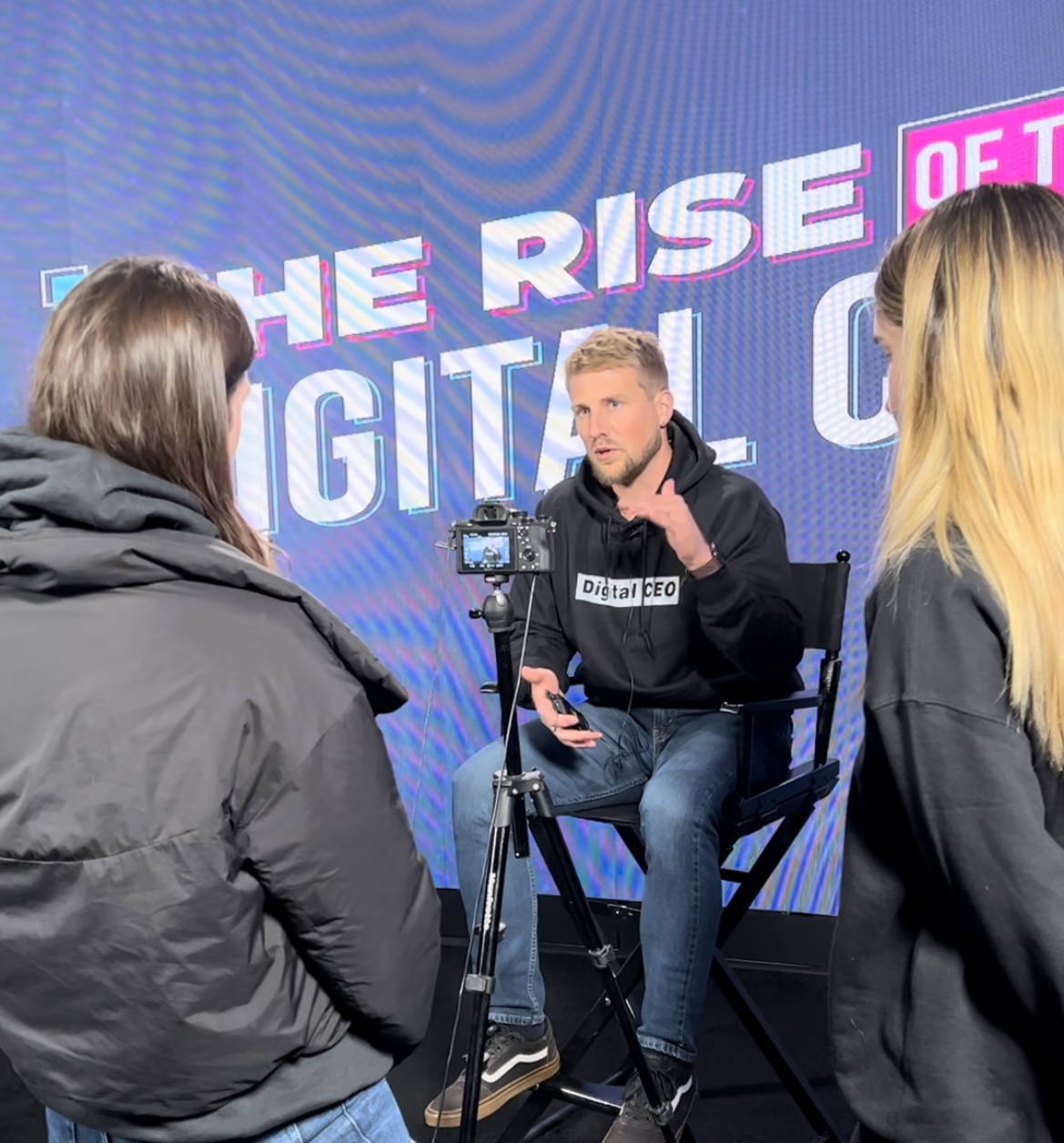 Man speaking into microphone during interview, two women in foreground, digital screen background with text, studio setting.
