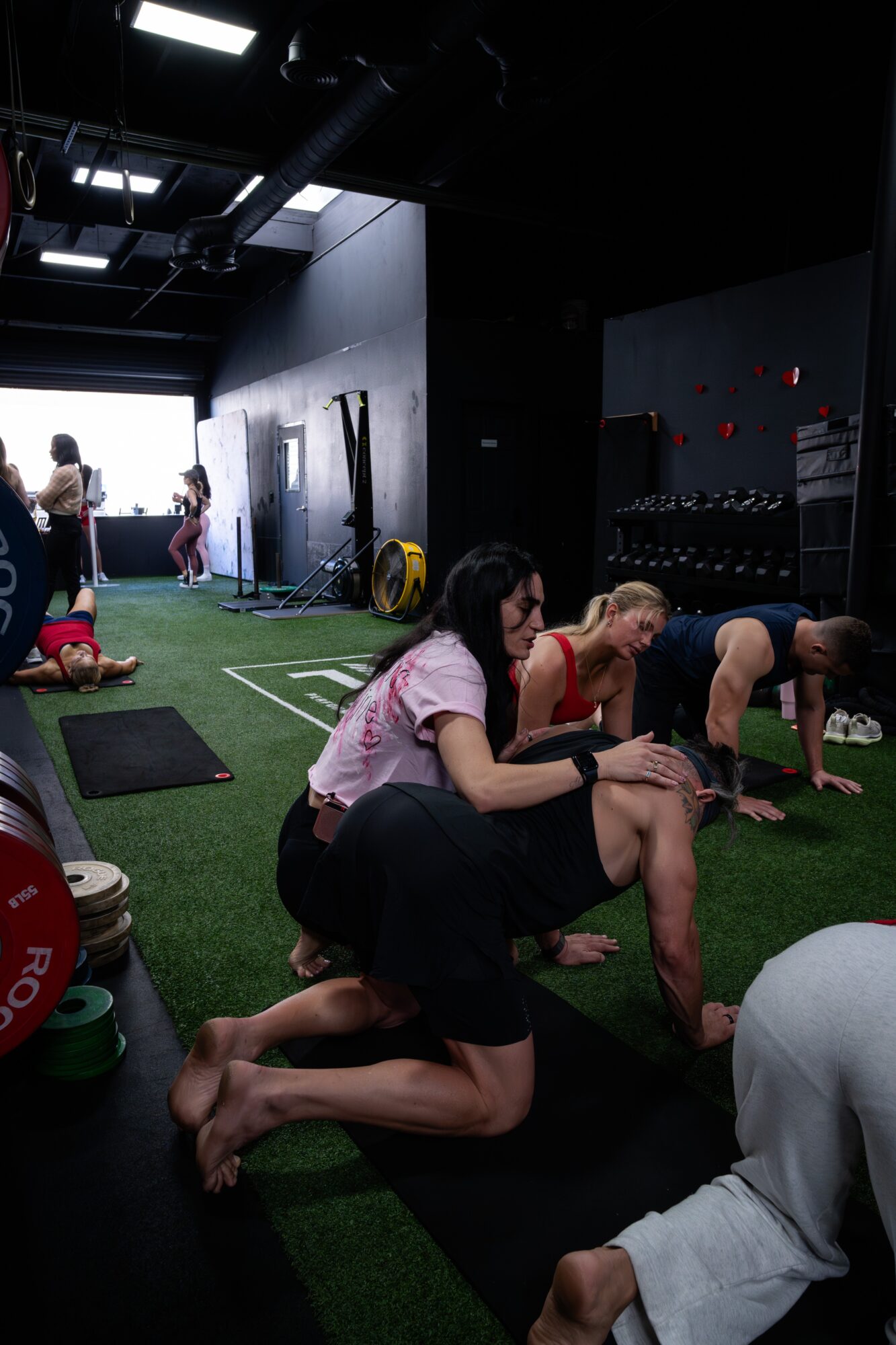People exercising in a gym with black walls, green turf, and equipment, some in plank positions, others standing or sitting.