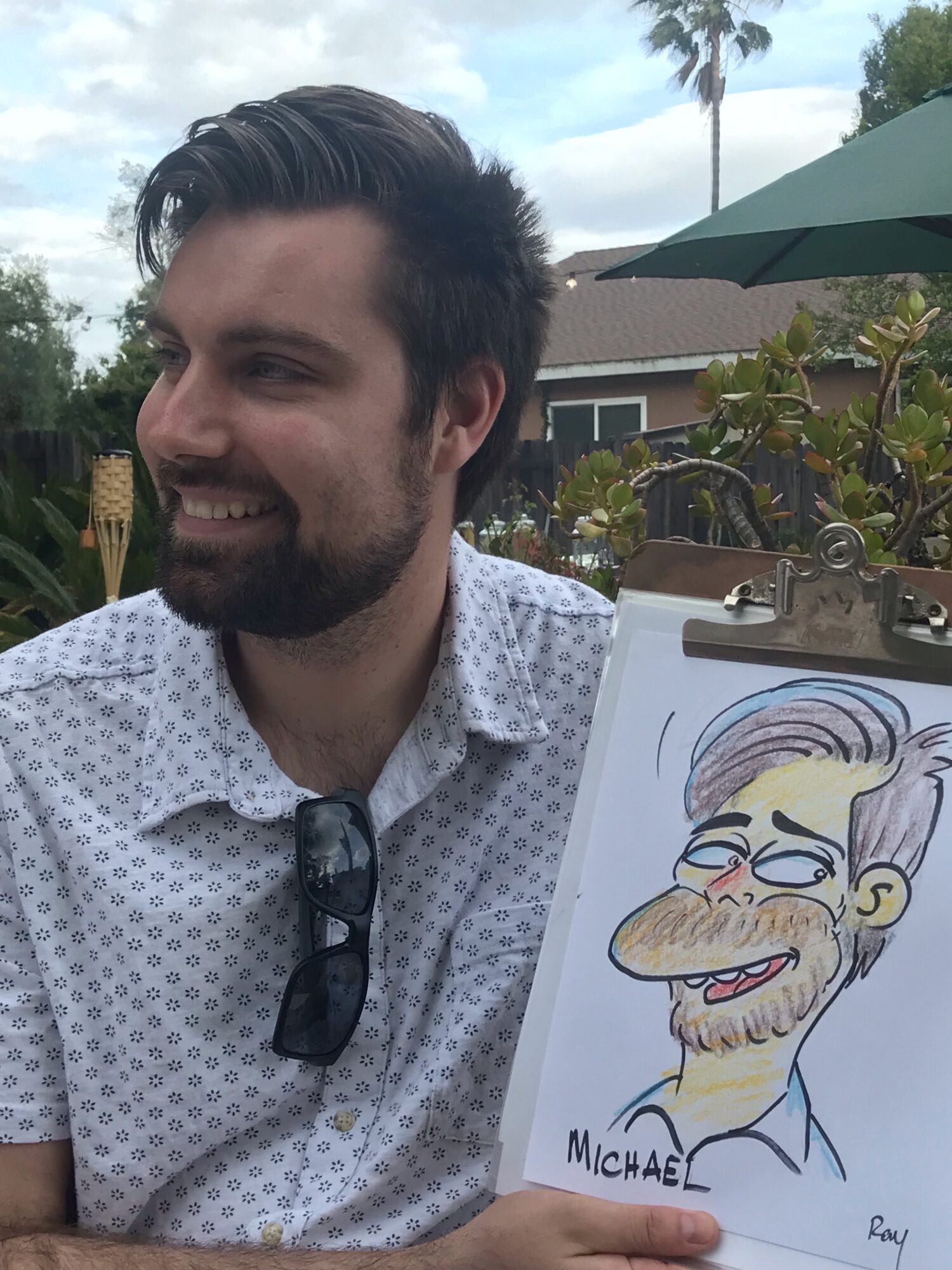 Man with dark hair and beard smiling outdoors, wearing a patterned shirt and sunglasses, with a cartoon drawing of a bearded man on a clipboard.