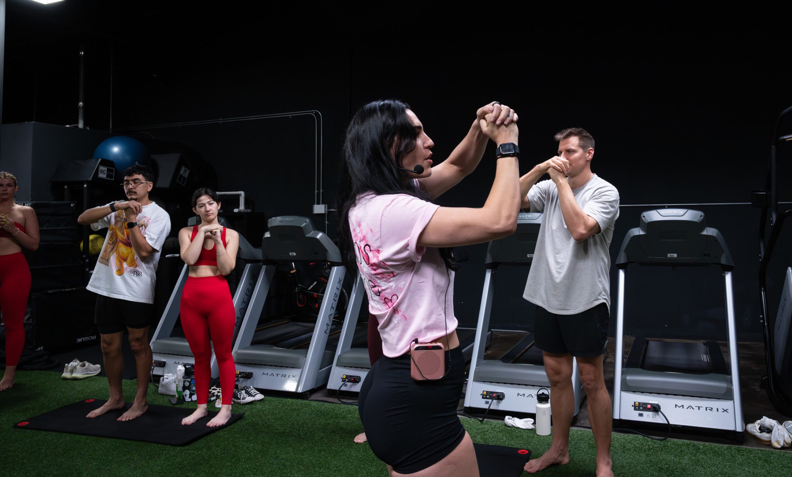 Group of people in a gym, some using treadmills, others standing and talking, with a woman in the foreground taking a photo.