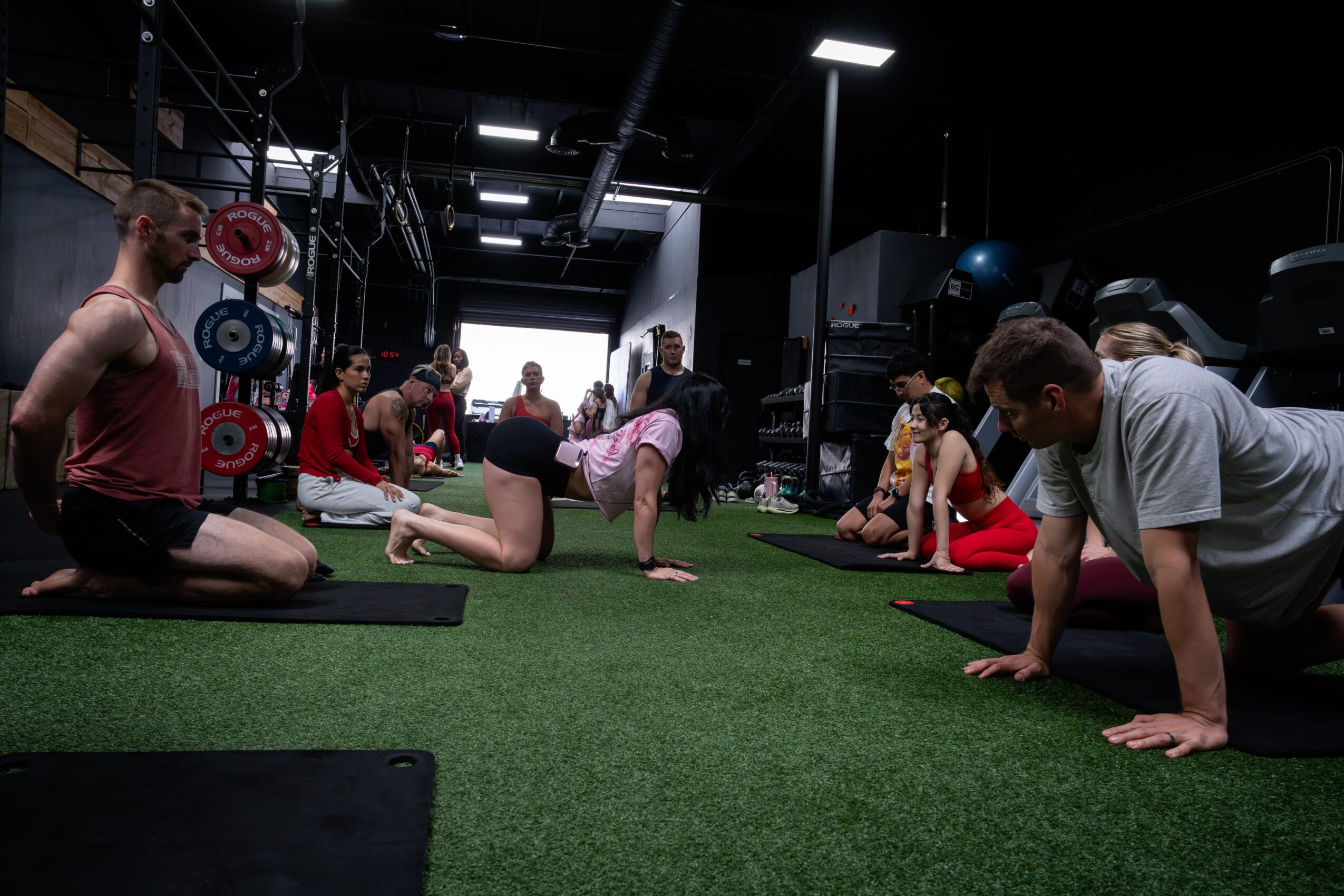 People participating in a fitness class on a green mat in a gym, with weights and equipment in the background.