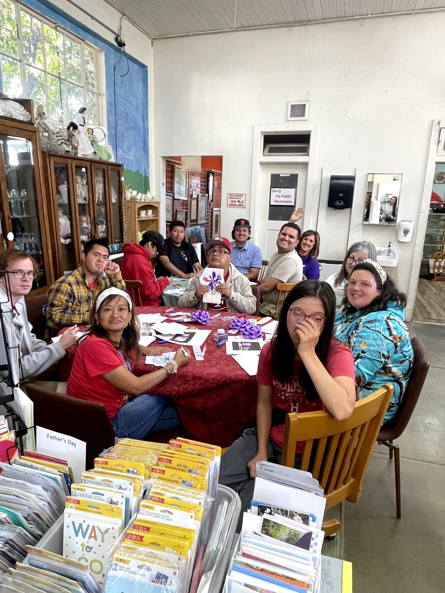 Group of people sitting around a table in a room with bookshelves and a window, smiling and engaging in activities.