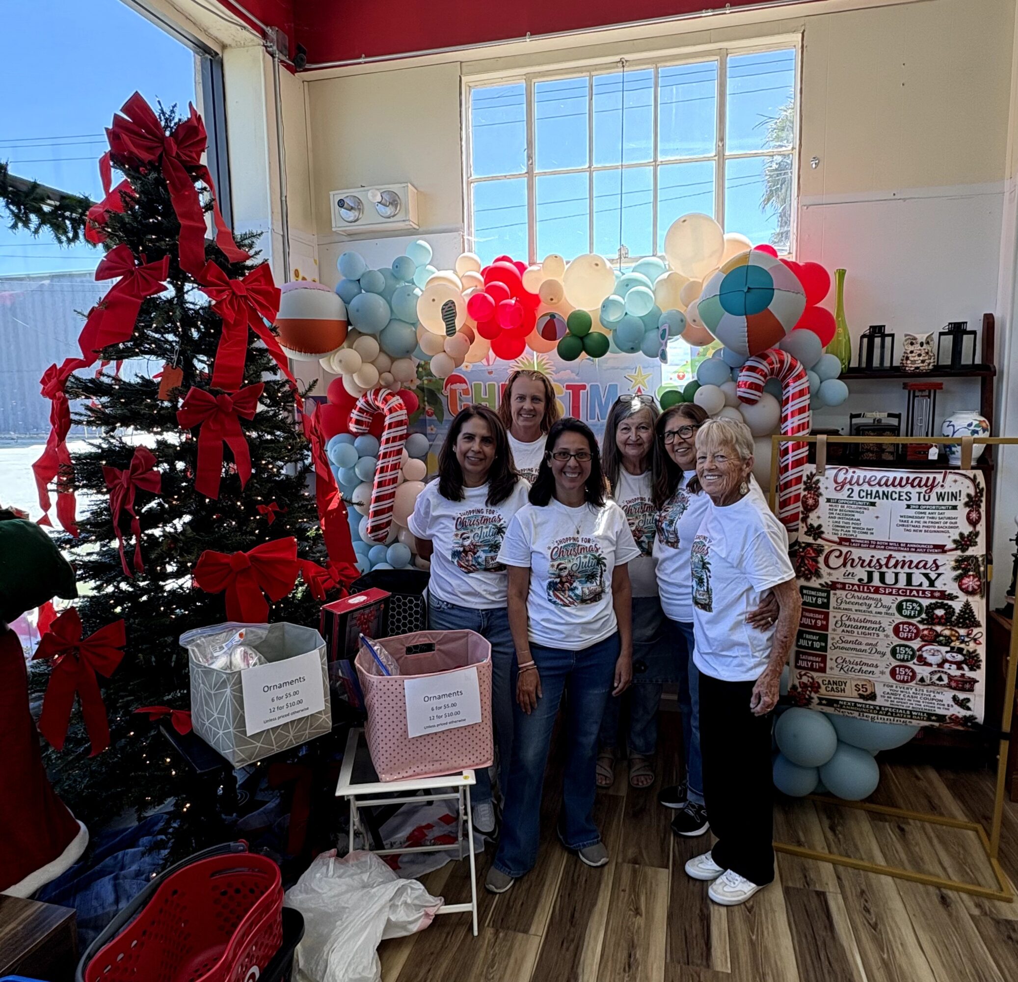 Group of five people standing indoors with Christmas decorations, balloons, and a tree, smiling at the camera.