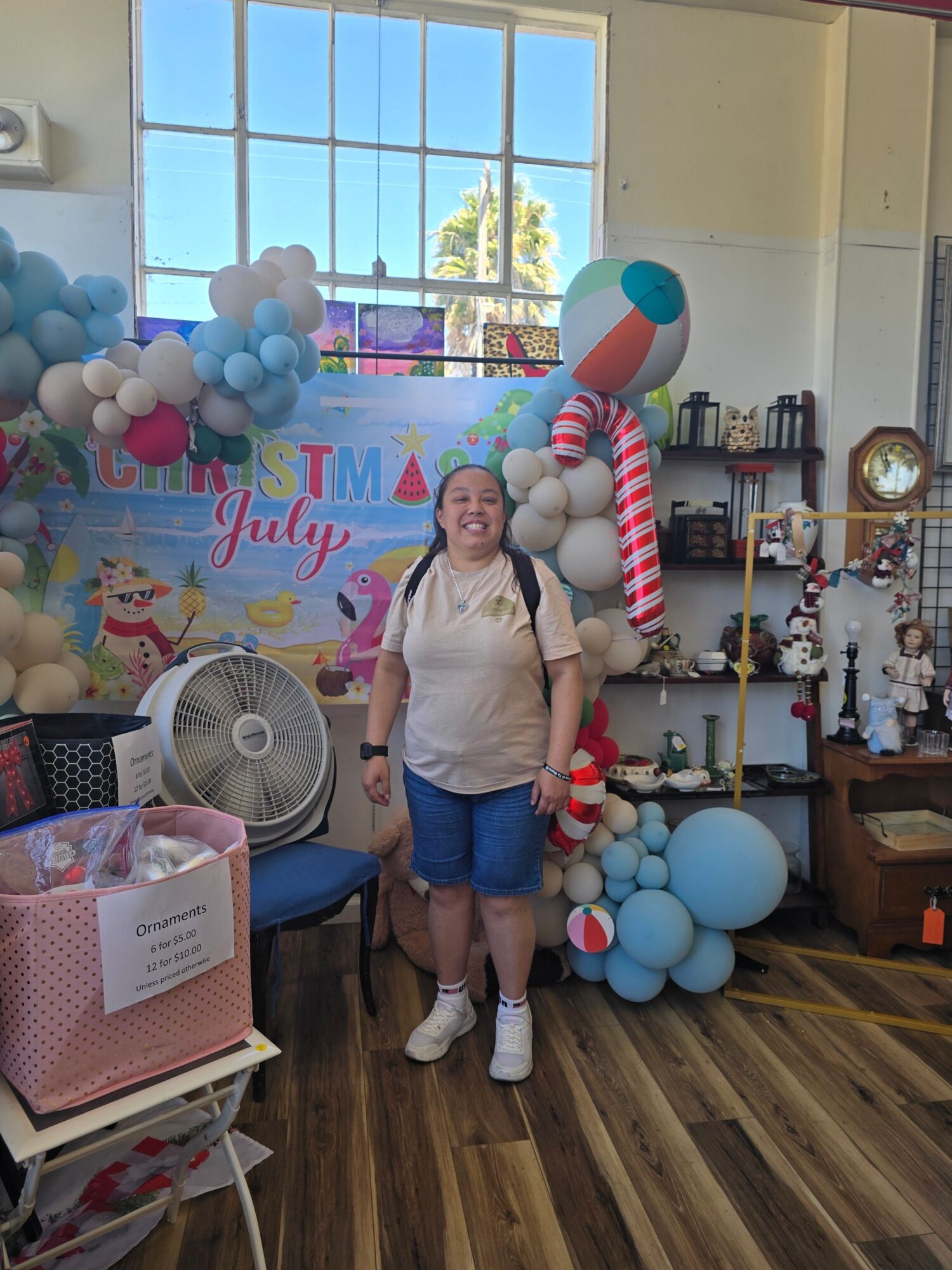 Person standing in decorated room with balloons, a banner, and a large striped candy prop, near a window and bookshelf.