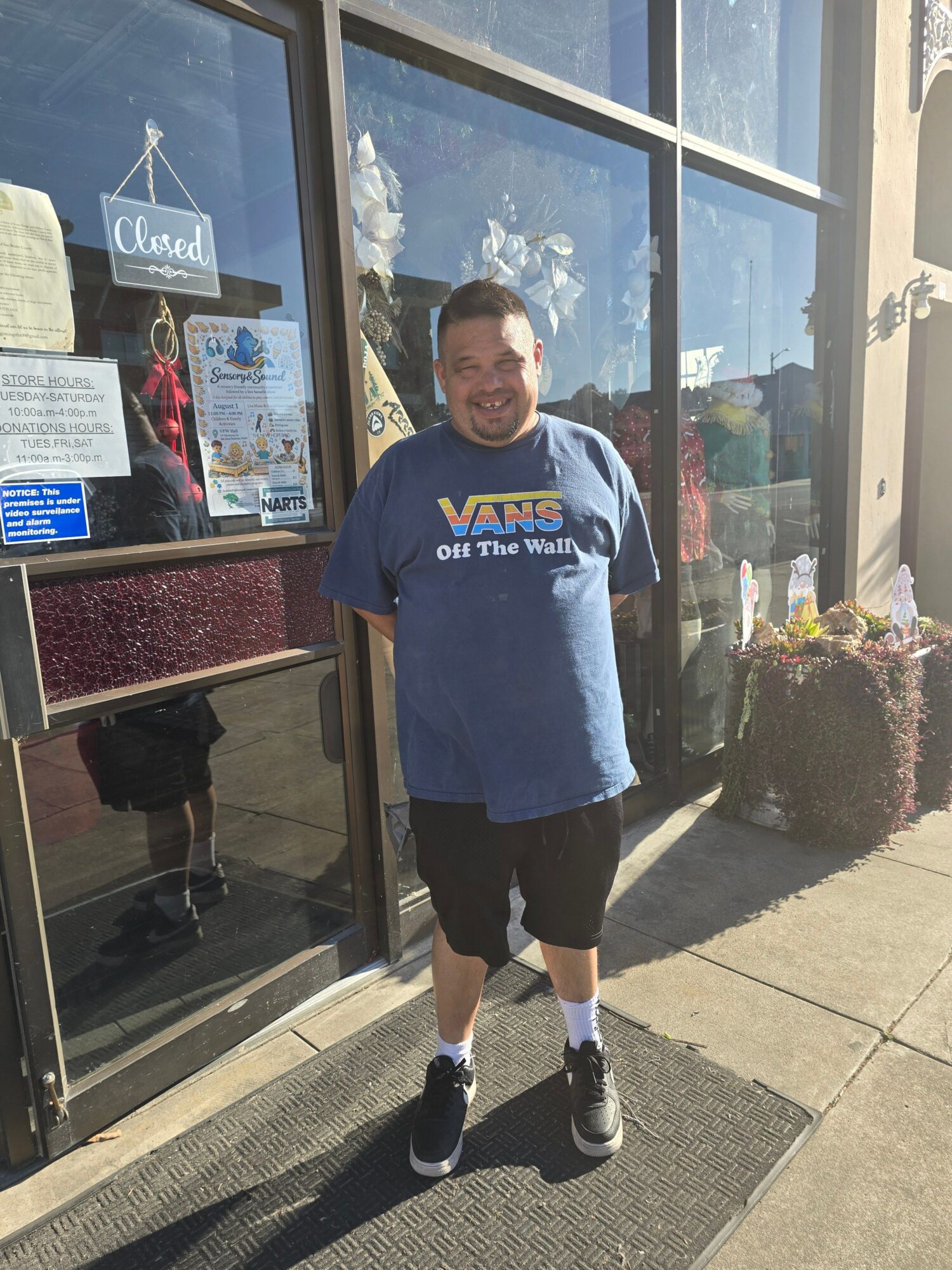 Person standing outside a glass storefront, wearing a blue sweatshirt and black shorts, with plants and sidewalk nearby.