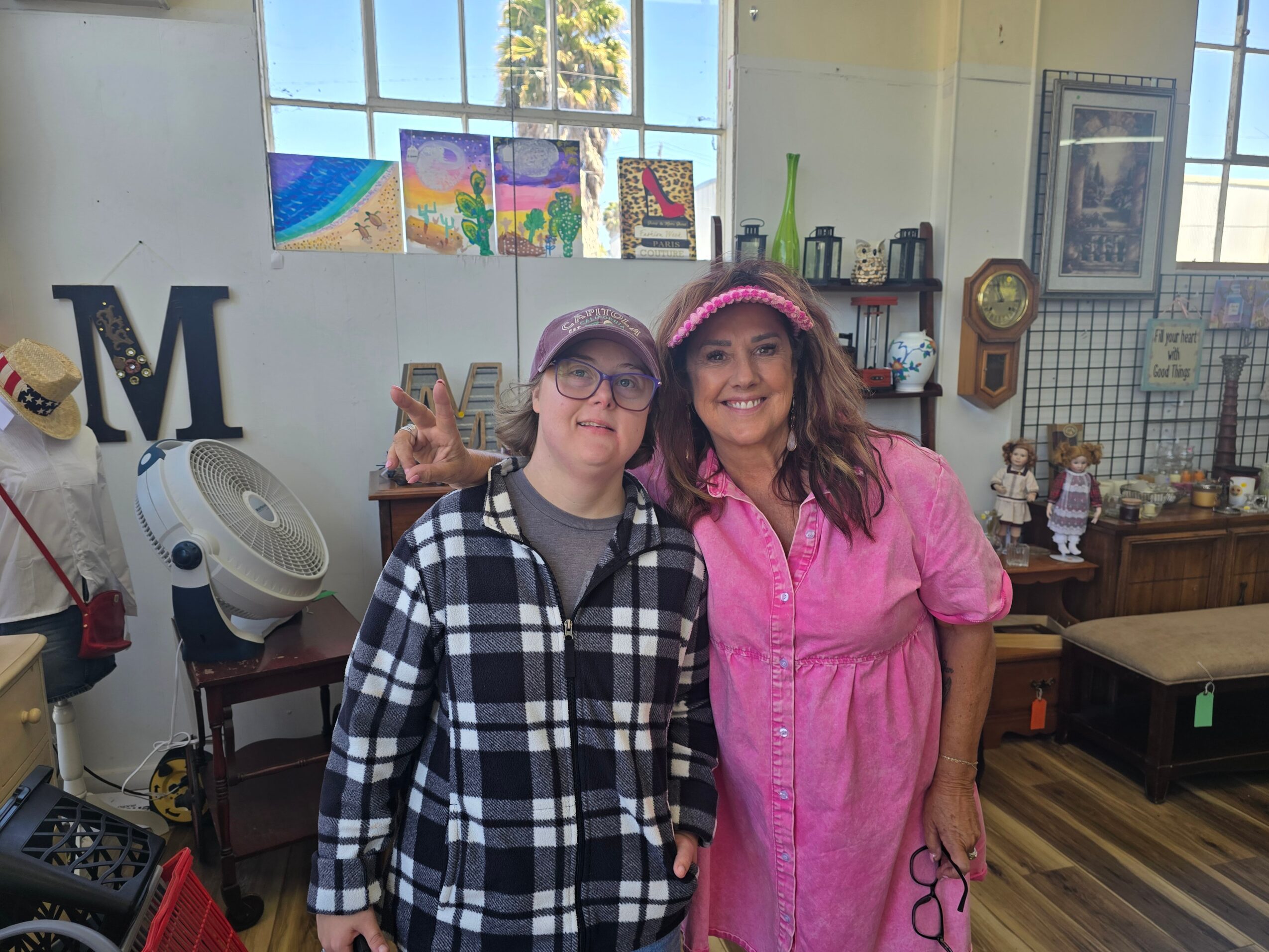 Two women standing indoors, smiling, with colorful artwork and shelves in the background.