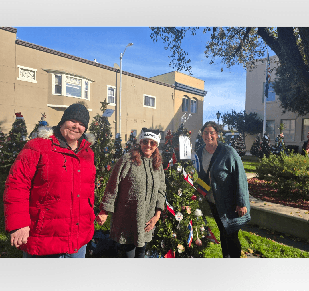 Three women stand outdoors near a decorated Christmas tree, with buildings and trees in the background.