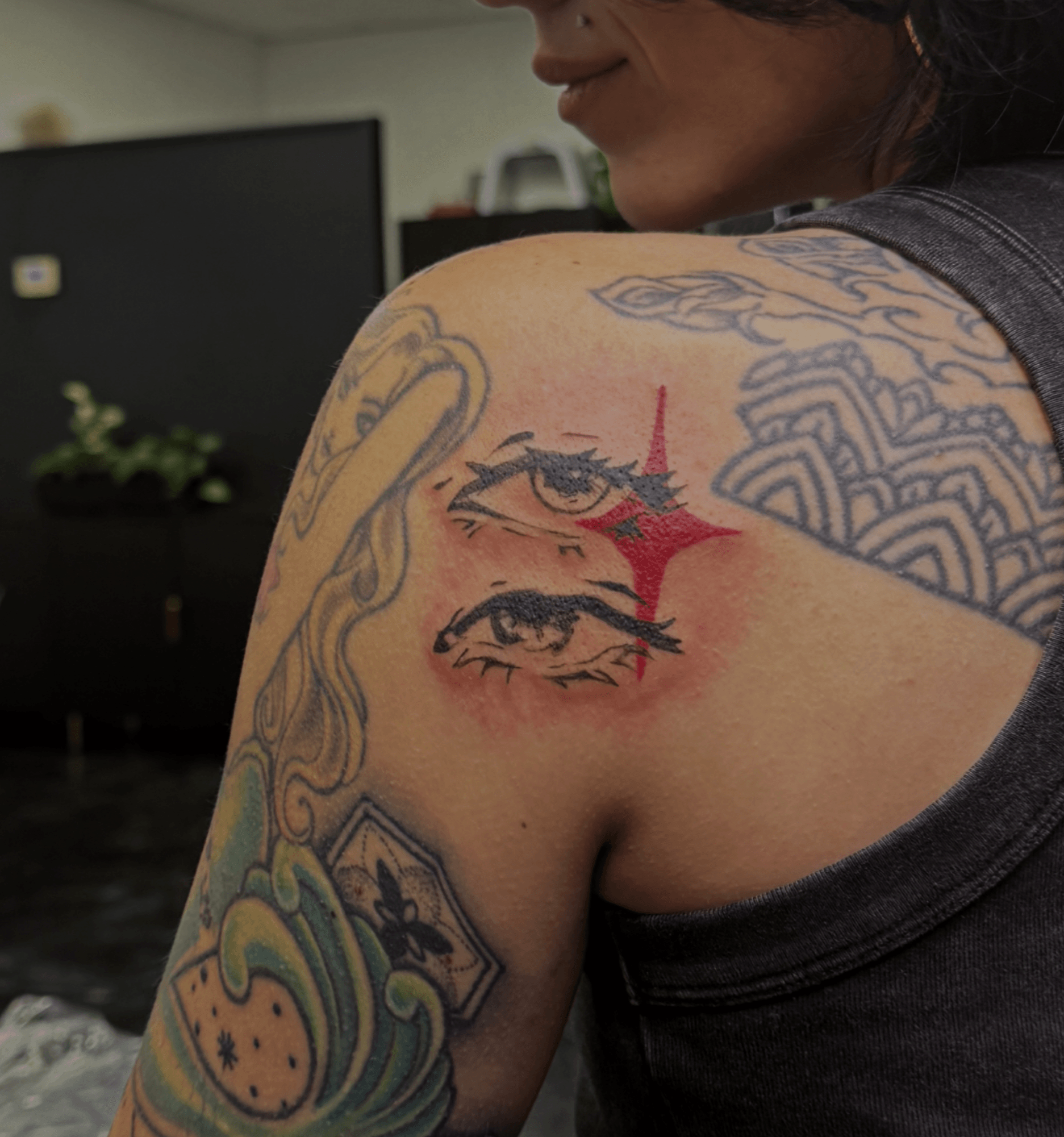 Close-up of a person's shoulder with colorful tattoos, including eyes, a lightning bolt, and abstract patterns.