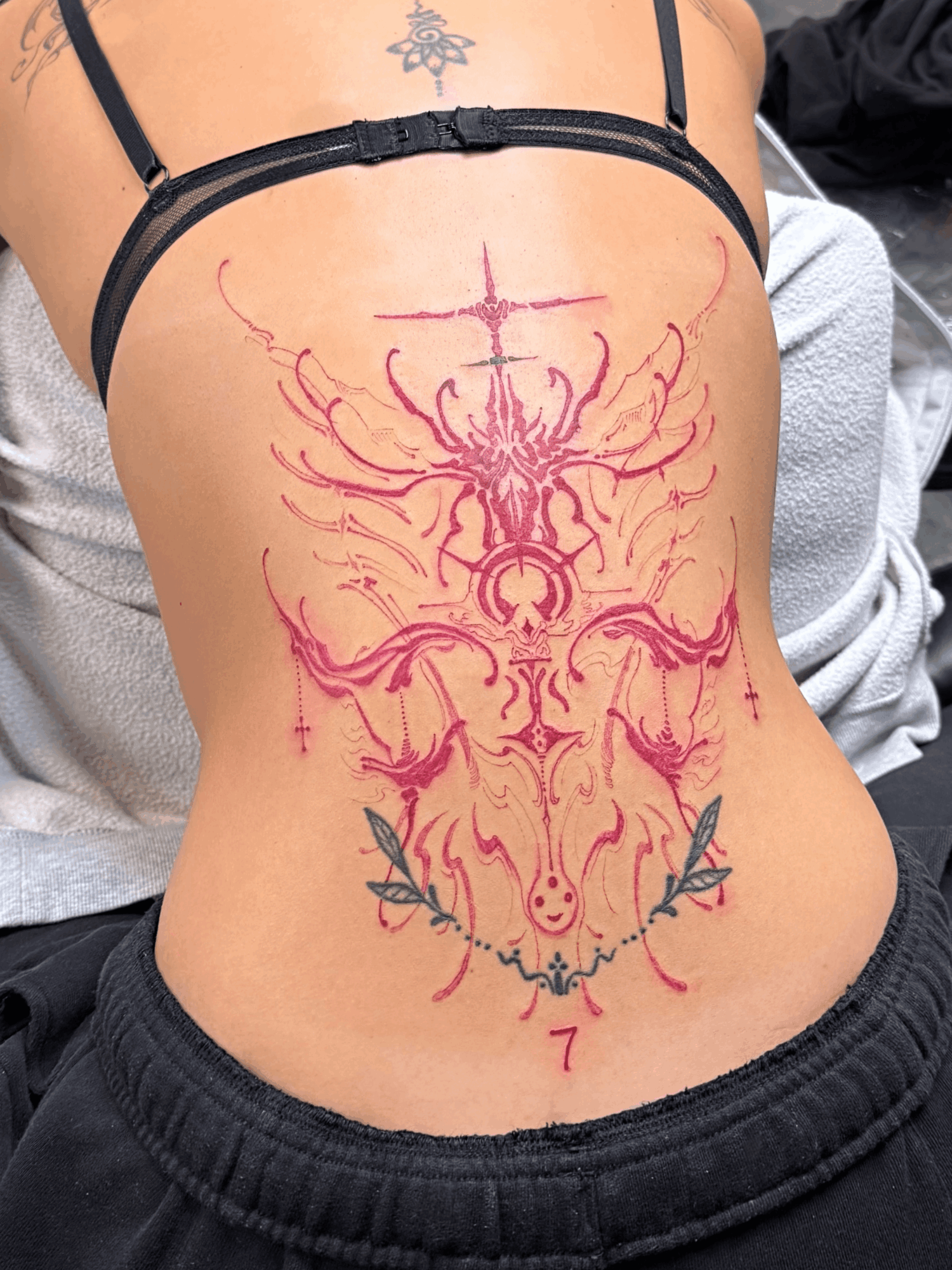 Colorful tattoo on person's back featuring a symmetrical design with a central skull, horns, and decorative elements, in pink, purple, and green.