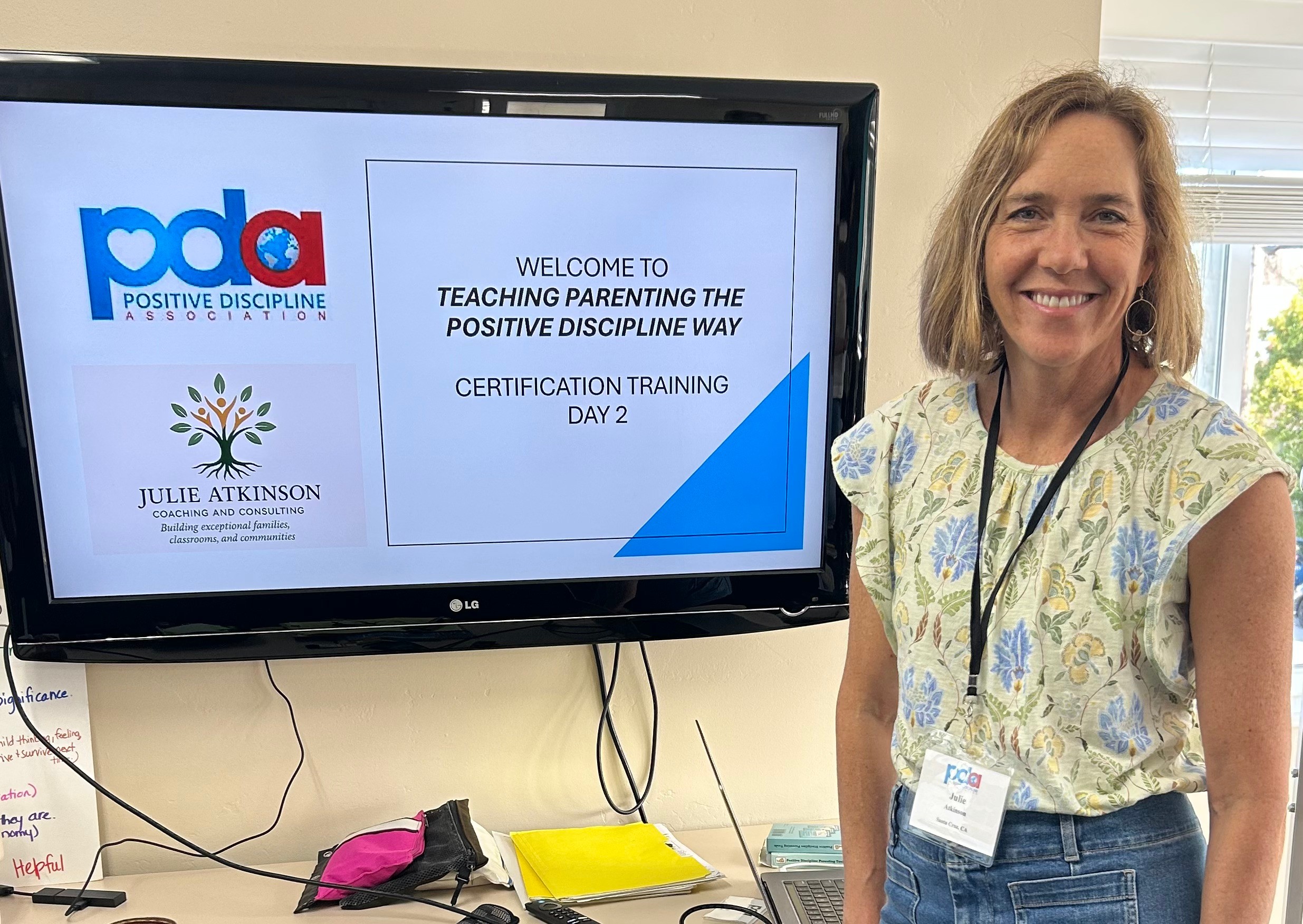 Woman smiling standing next to a screen displaying a presentation titled 'Teaching Parenting the Positive Discipline Way' at a training event.