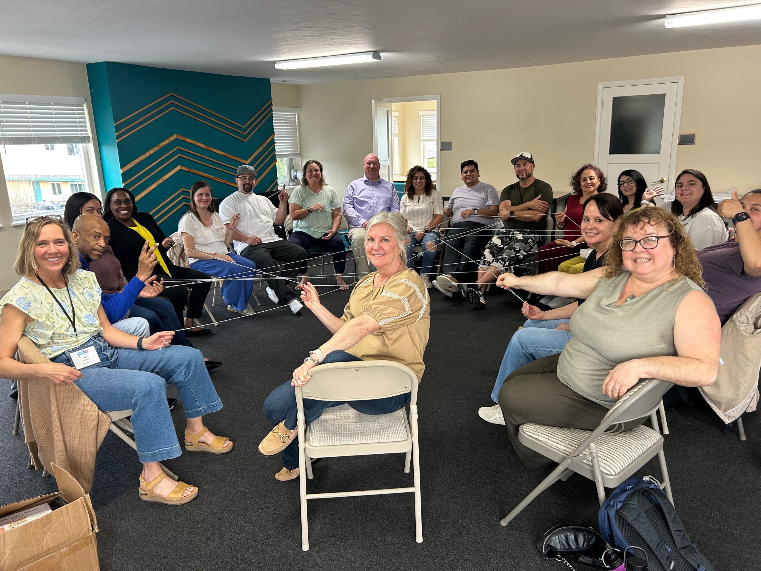 Group of people sitting in a room, holding strings connected to a central person, smiling and engaging in activity.