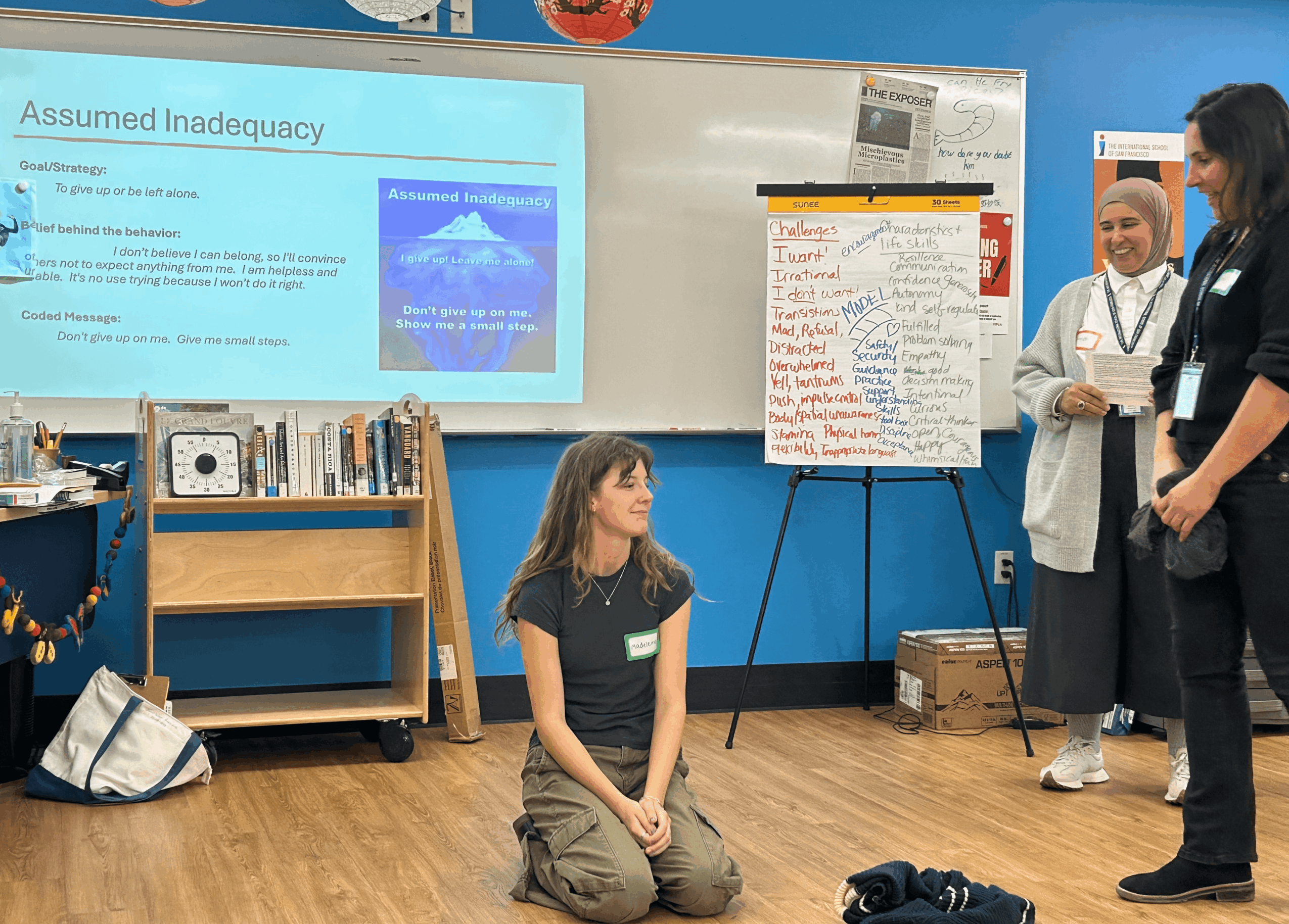 Girl kneeling on the floor in a classroom, with two women standing nearby, a whiteboard, and a presentation slide on the wall.