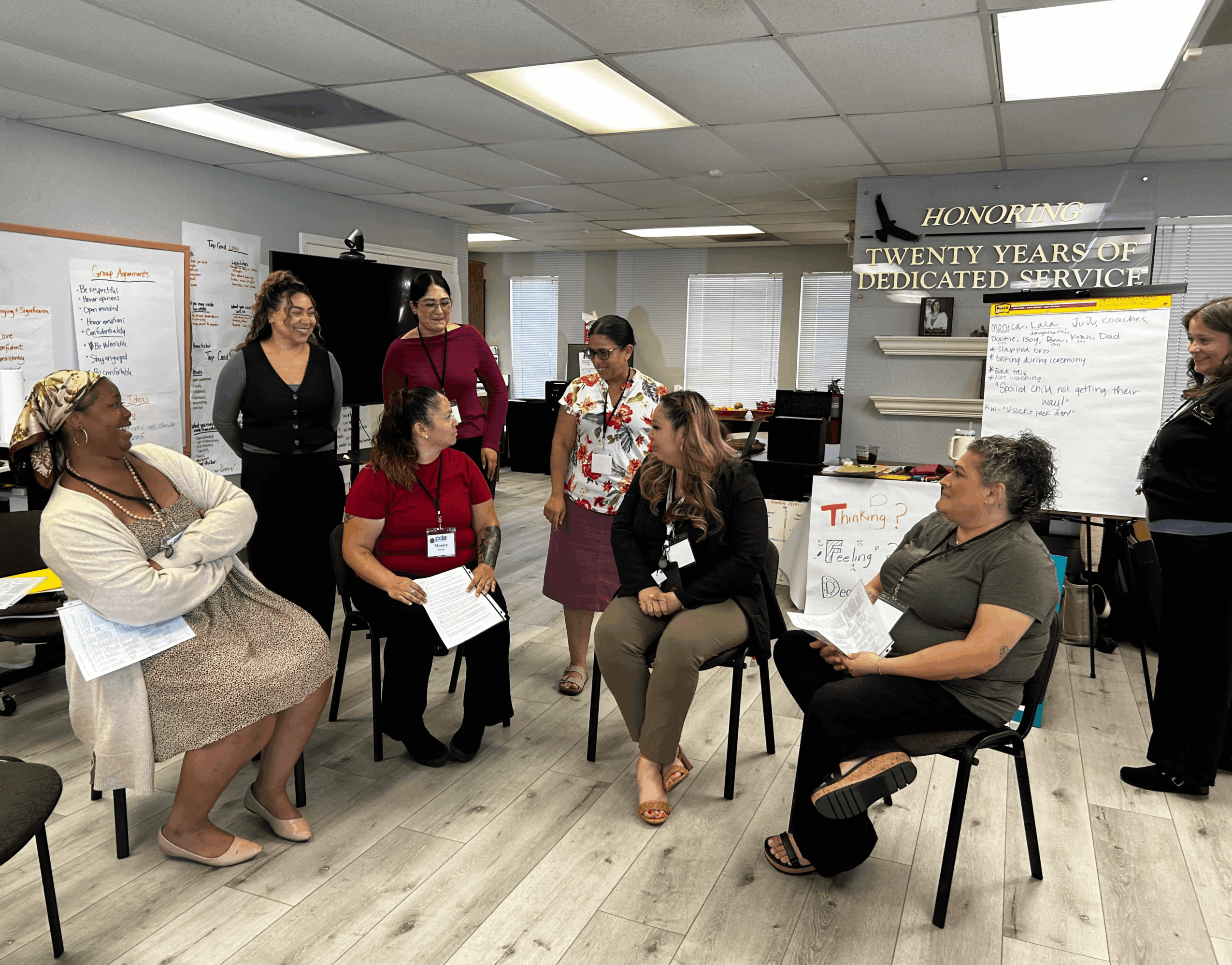 Group of women in a room with chairs, some seated and some standing, engaged in conversation, with posters and a sign in the background.