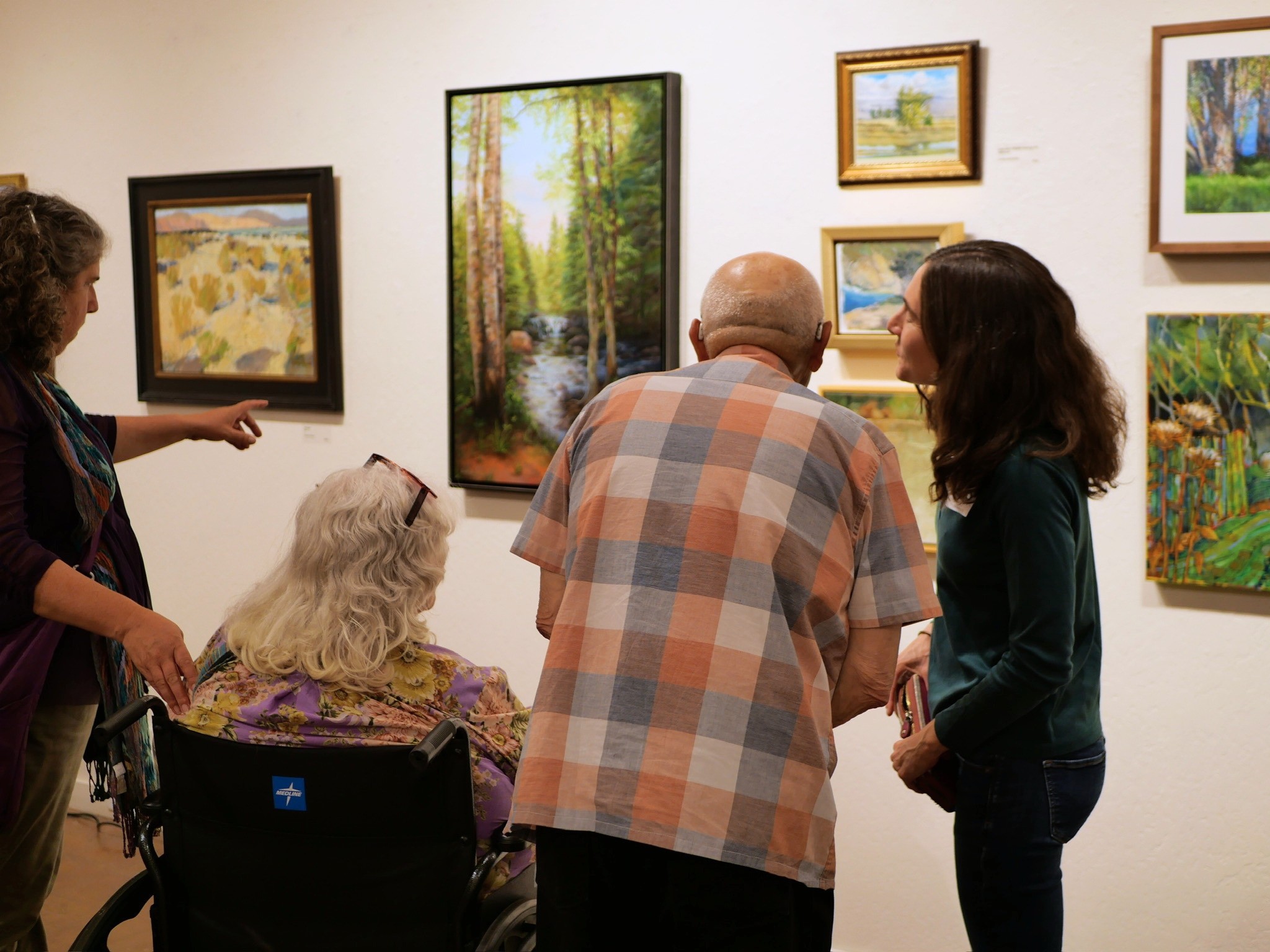 Group of four people viewing paintings in an art gallery, with one person in a wheelchair, discussing artwork.