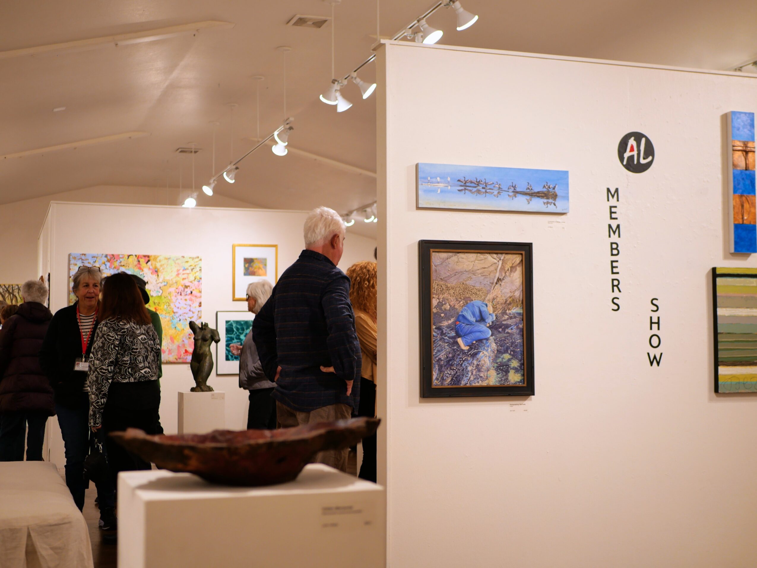 People viewing artwork in an art gallery with white walls and colorful paintings, some people are talking and observing art.