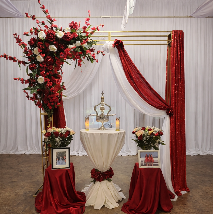 Decorative setup with red and white flowers, draped fabric, and candles, likely for a celebration or wedding, with a central table and floral arrangements.
