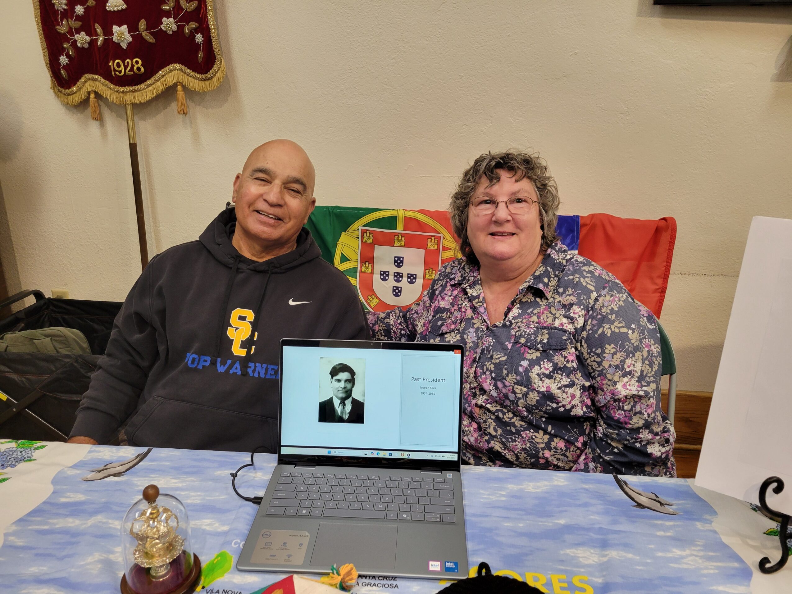 Two people sitting at a table with a laptop displaying a black-and-white photo, a flag, and a decorative banner in the background.