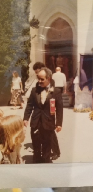 Person in formal attire standing outdoors near a building entrance, with other people in the background.