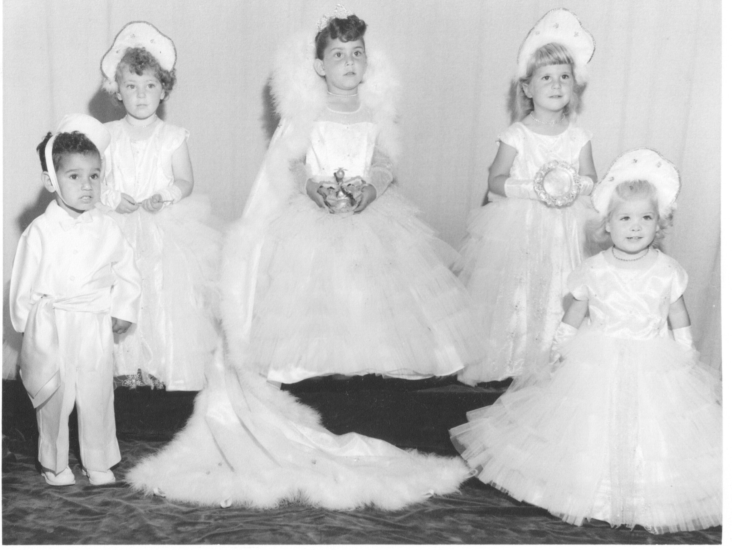 Group of six children dressed in formal attire, with three girls in dresses and two boys in suits, standing indoors.