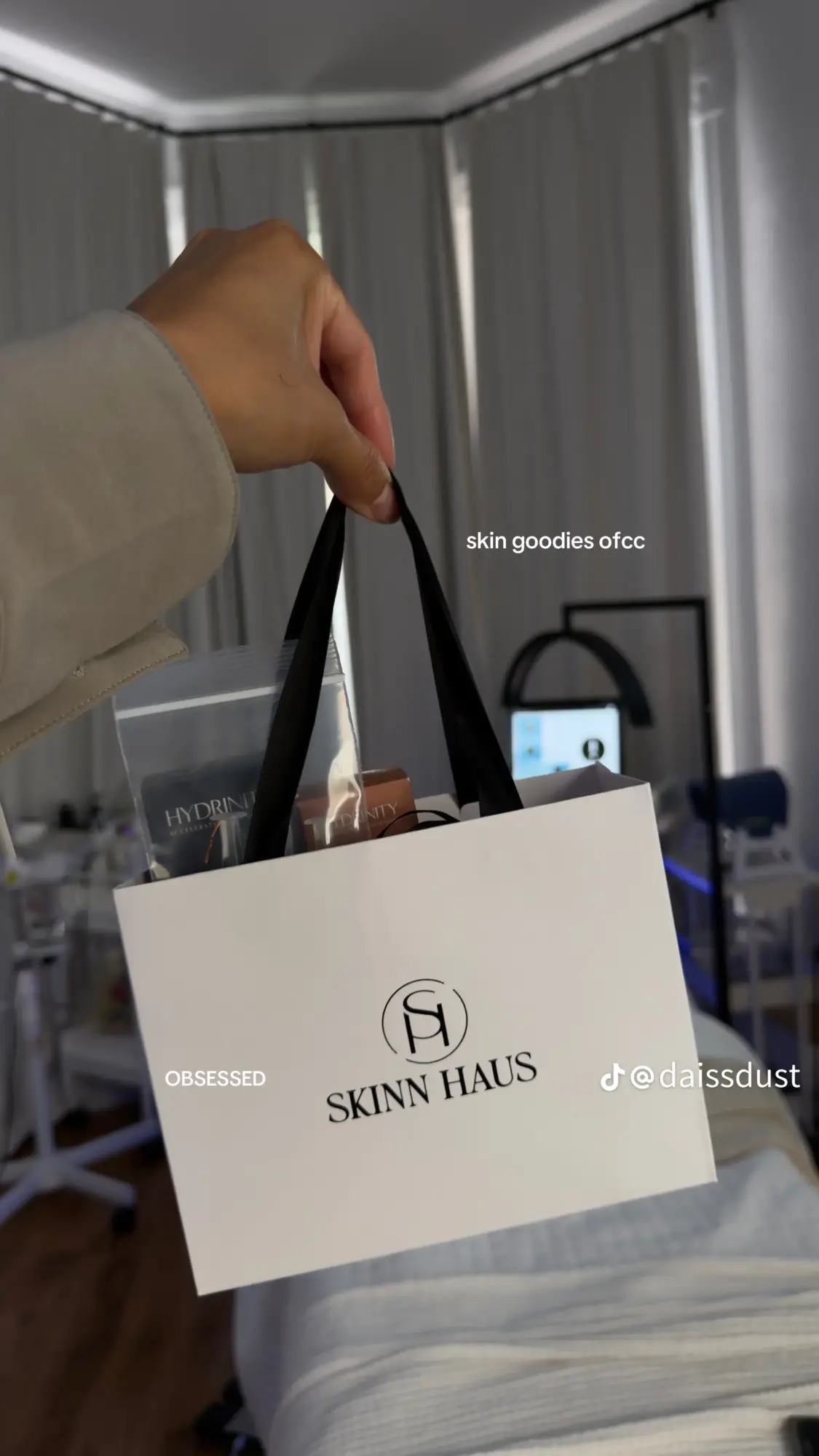 Person holding a white shopping bag with black handles in a room with curtains and a bed.