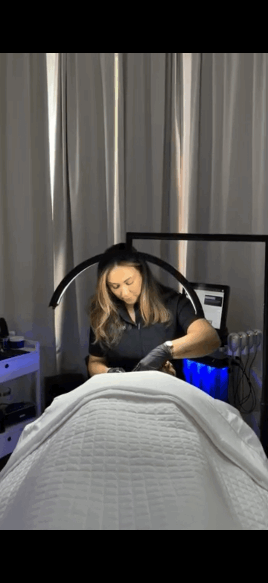 A woman in a medical setting with curtains and equipment, looking at her phone, viewed from the foot of a bed.