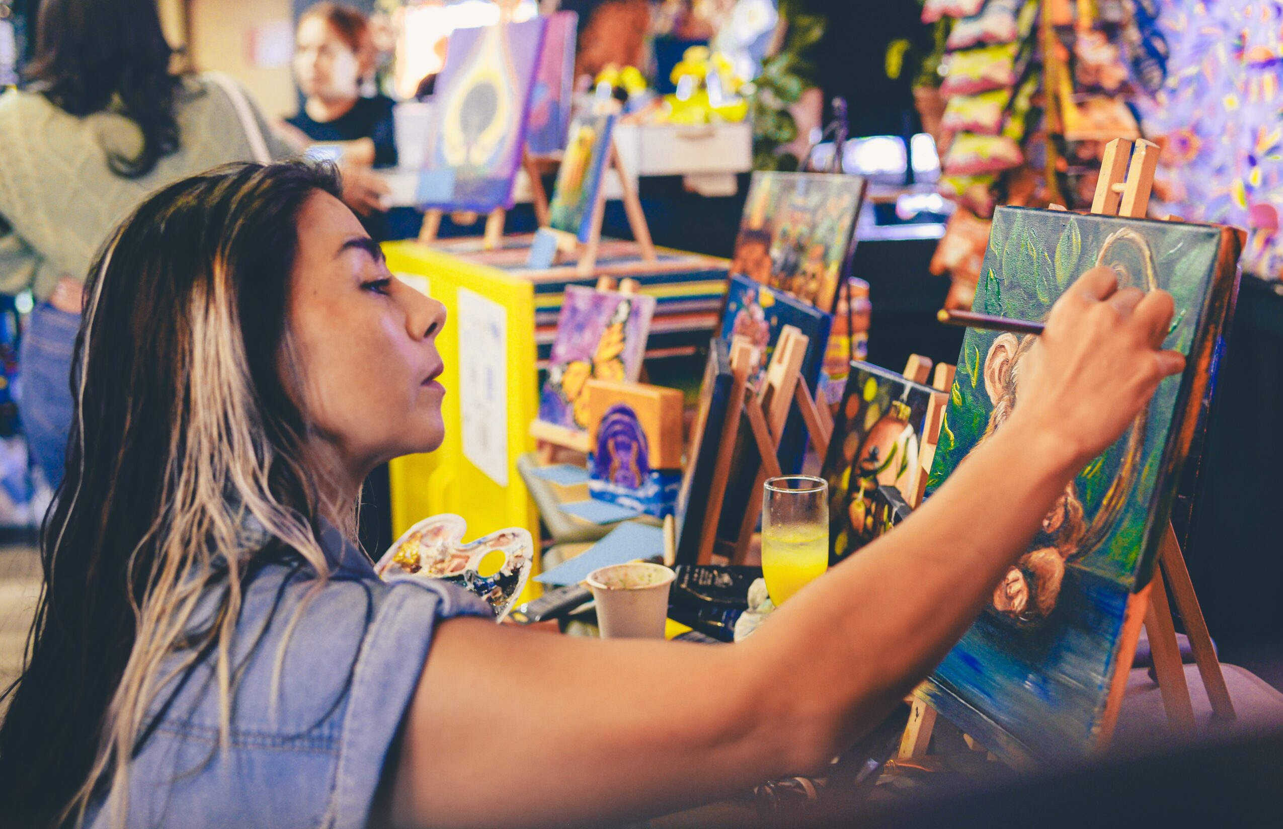 Woman painting on canvas at an art event with colorful paintings and people in background.