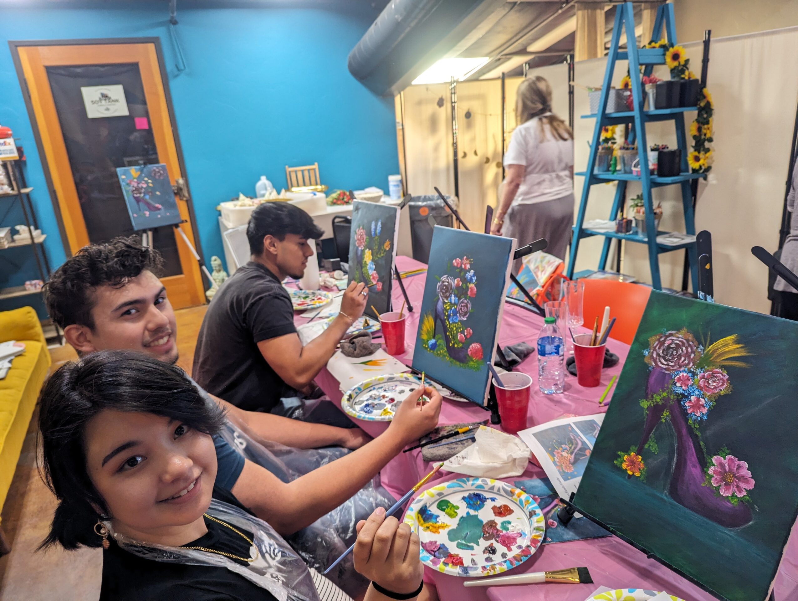 Children painting floral artwork at a table in an art studio, with a woman in the background and art supplies visible.