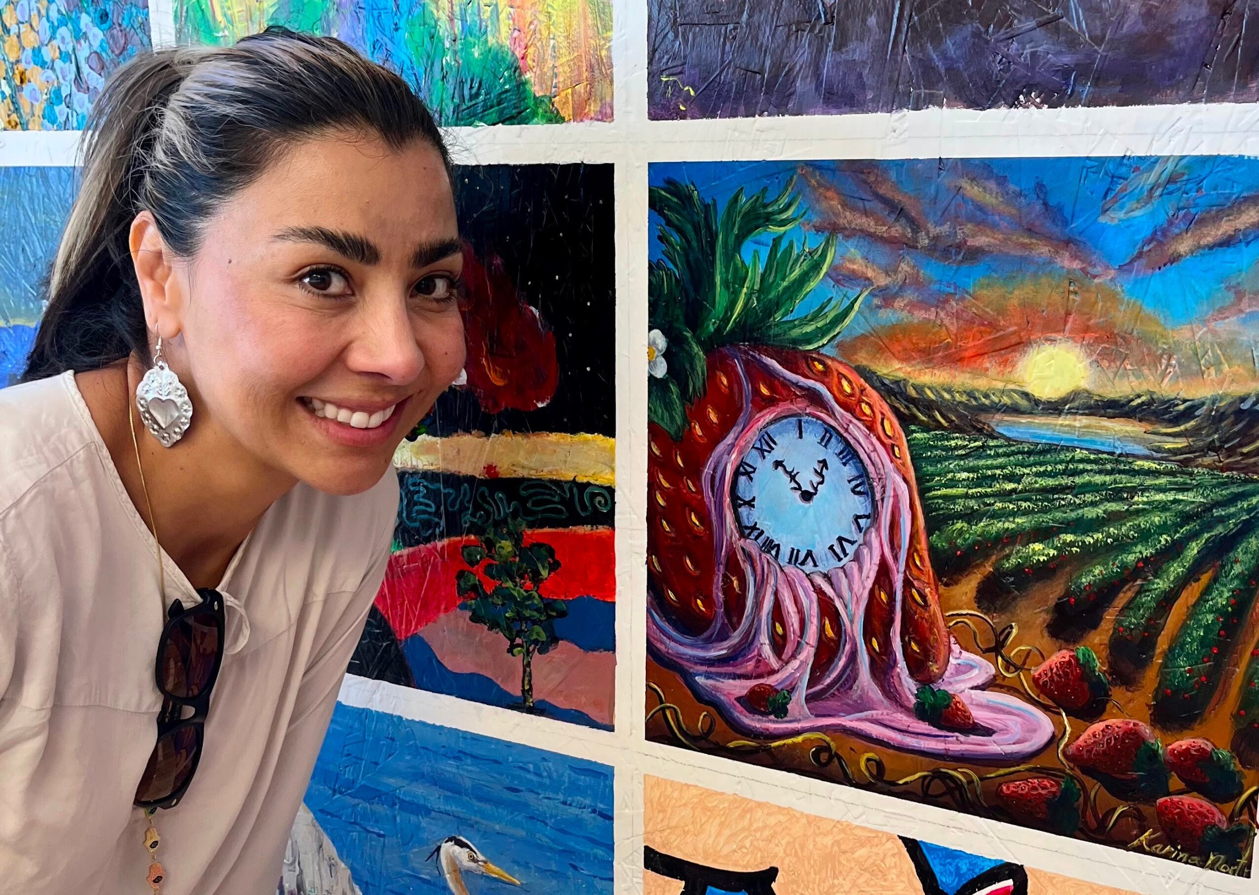 Smiling woman with dark hair and earrings stands next to colorful paintings, including a landscape with a clock and sunset.