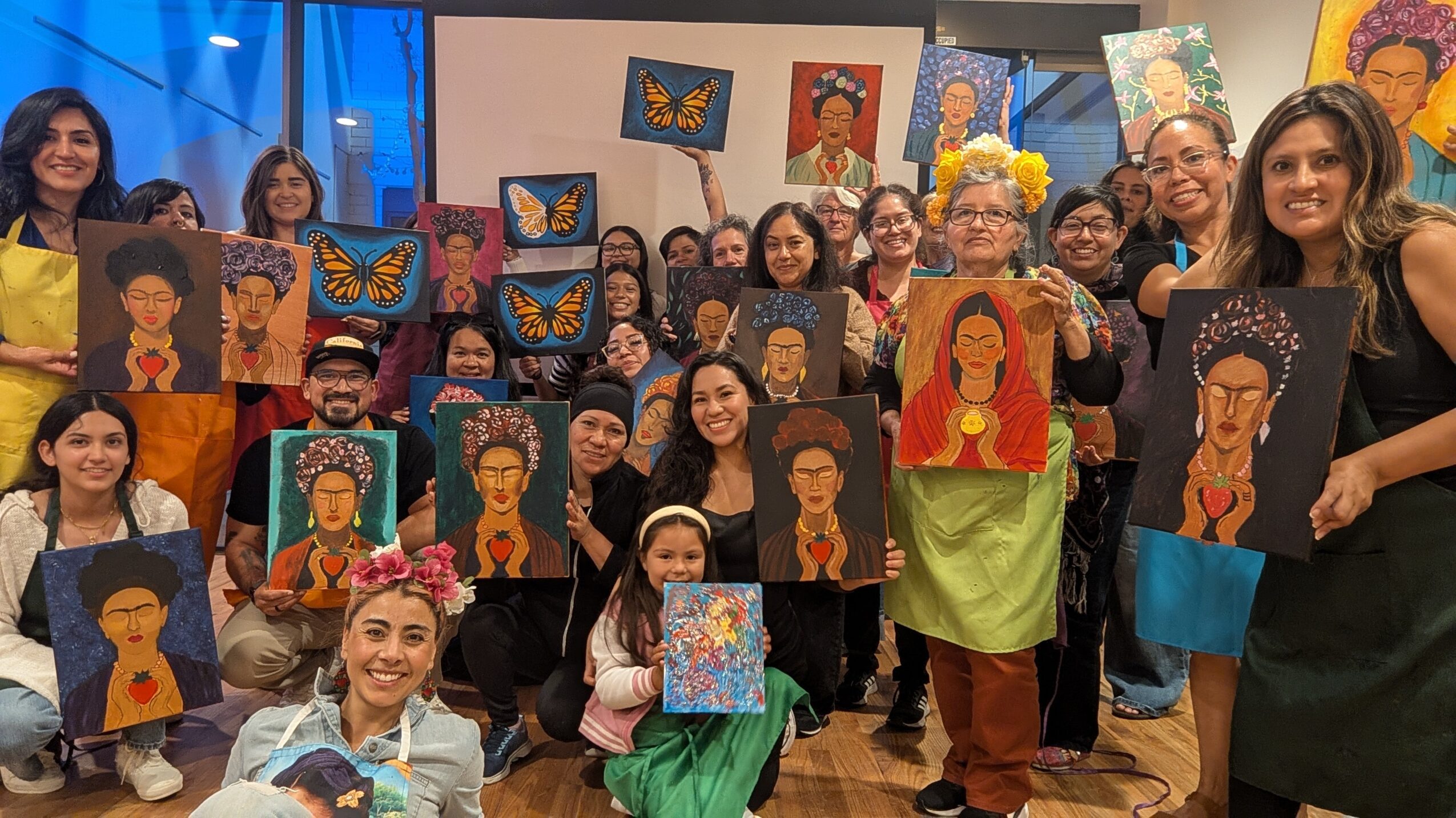 Group of people holding colorful portraits and artwork, smiling indoors with blue lighting in the background.