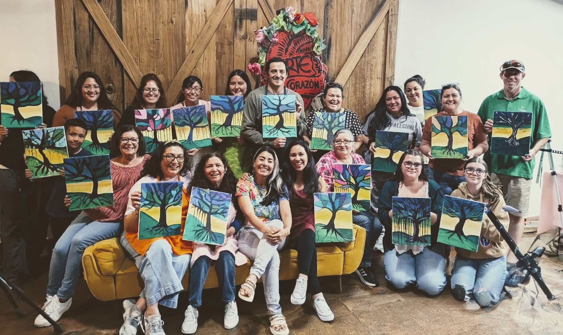 Group of people holding paintings, sitting and standing in front of a wooden wall with a floral decoration, smiling.