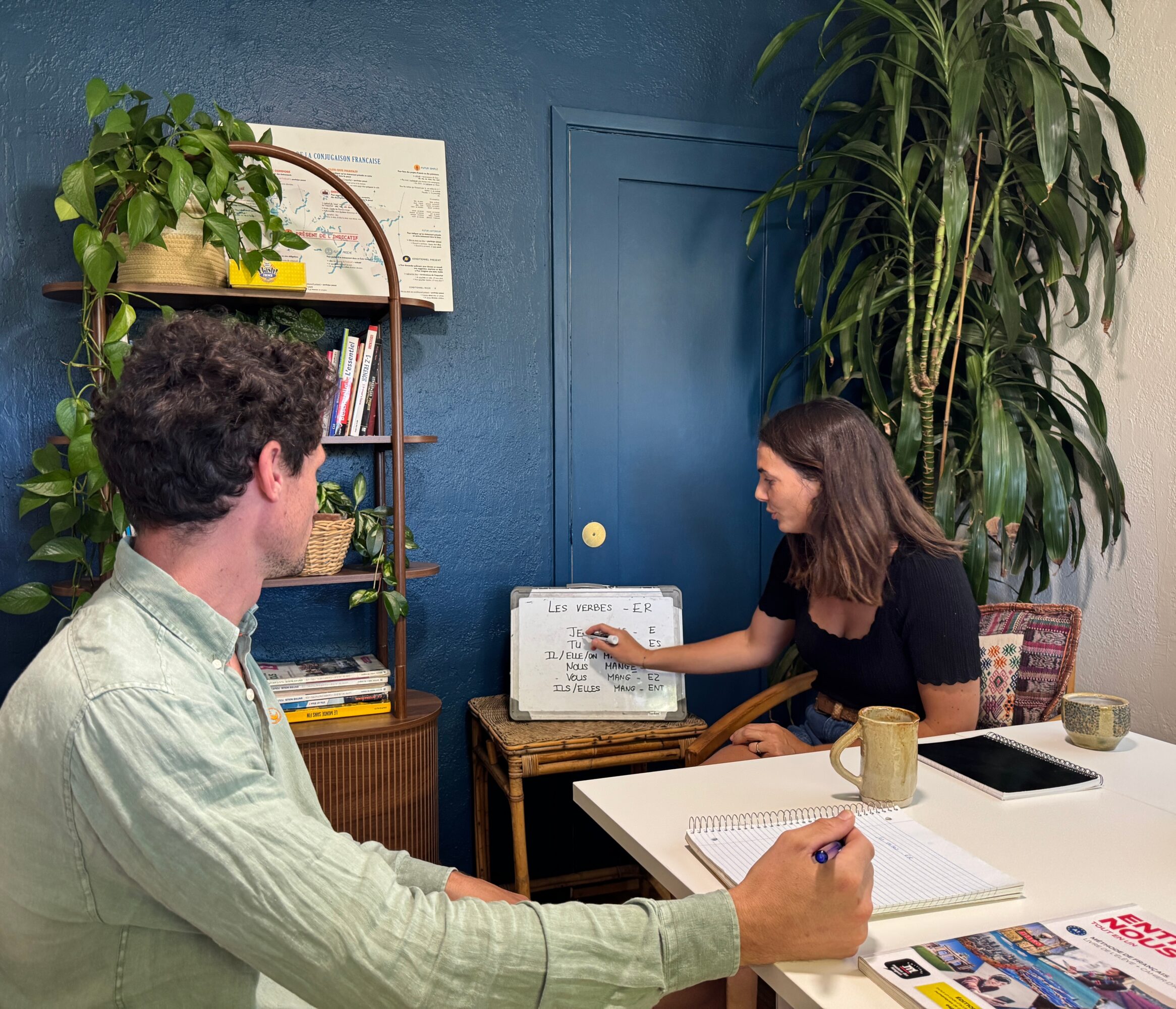 Two people in a room with blue walls, one sitting at a desk and the other pointing at a whiteboard.