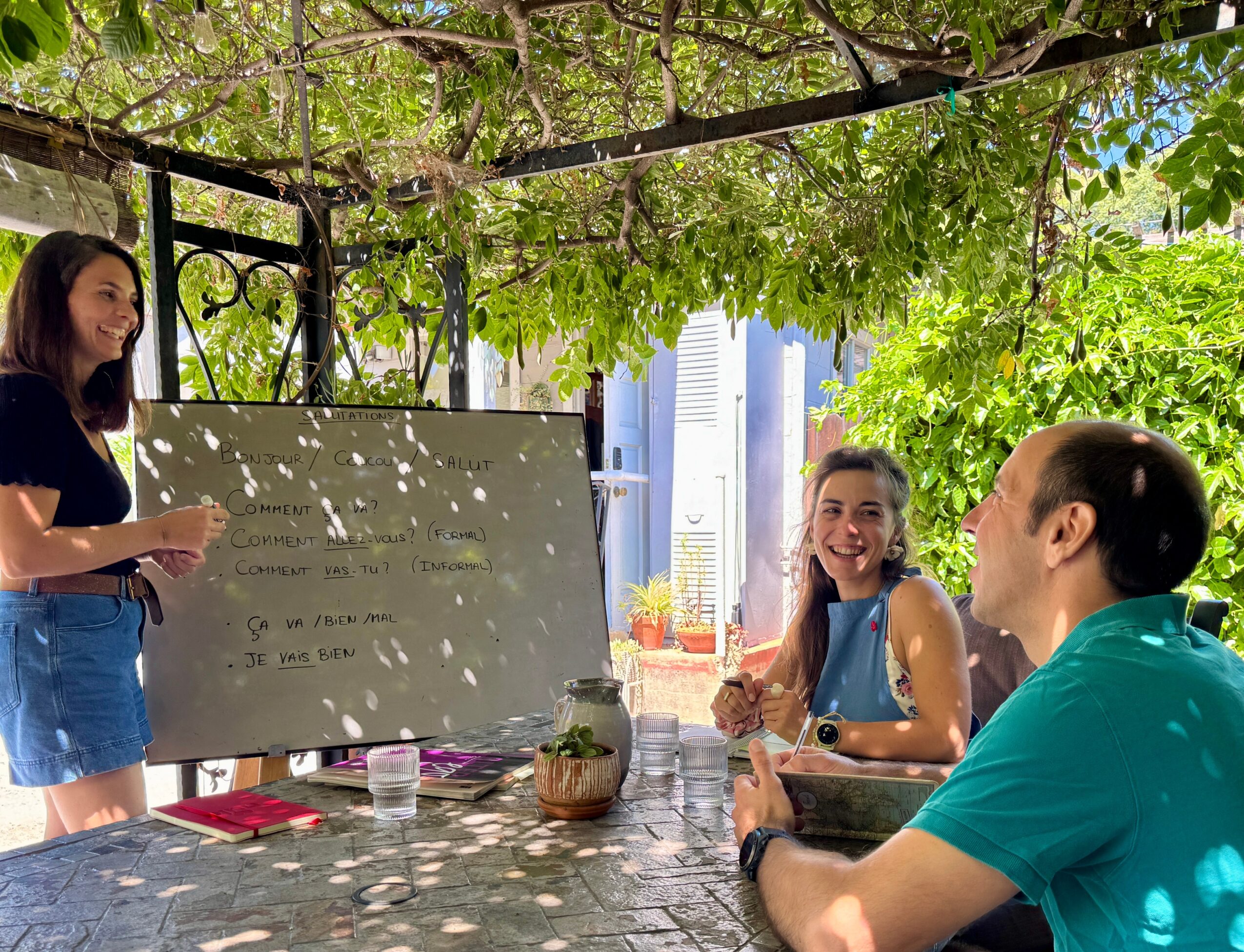 Three people sitting and standing around a table outdoors, with a whiteboard and greenery overhead.