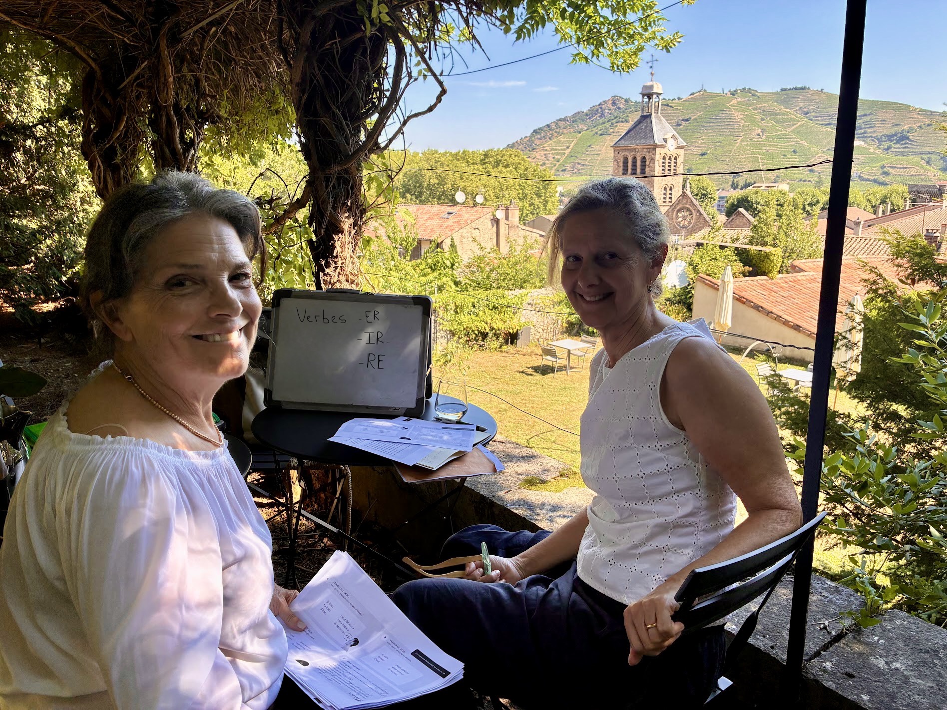 Two women sitting outdoors with a laptop, trees, and a village with hills in the background.