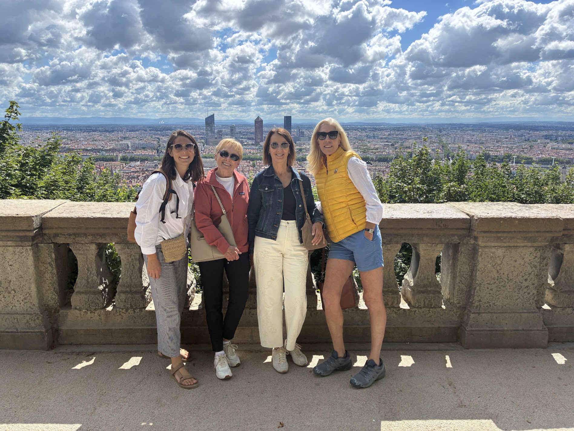 Four women stand together on a stone balcony with a city skyline and cloudy sky in the background.