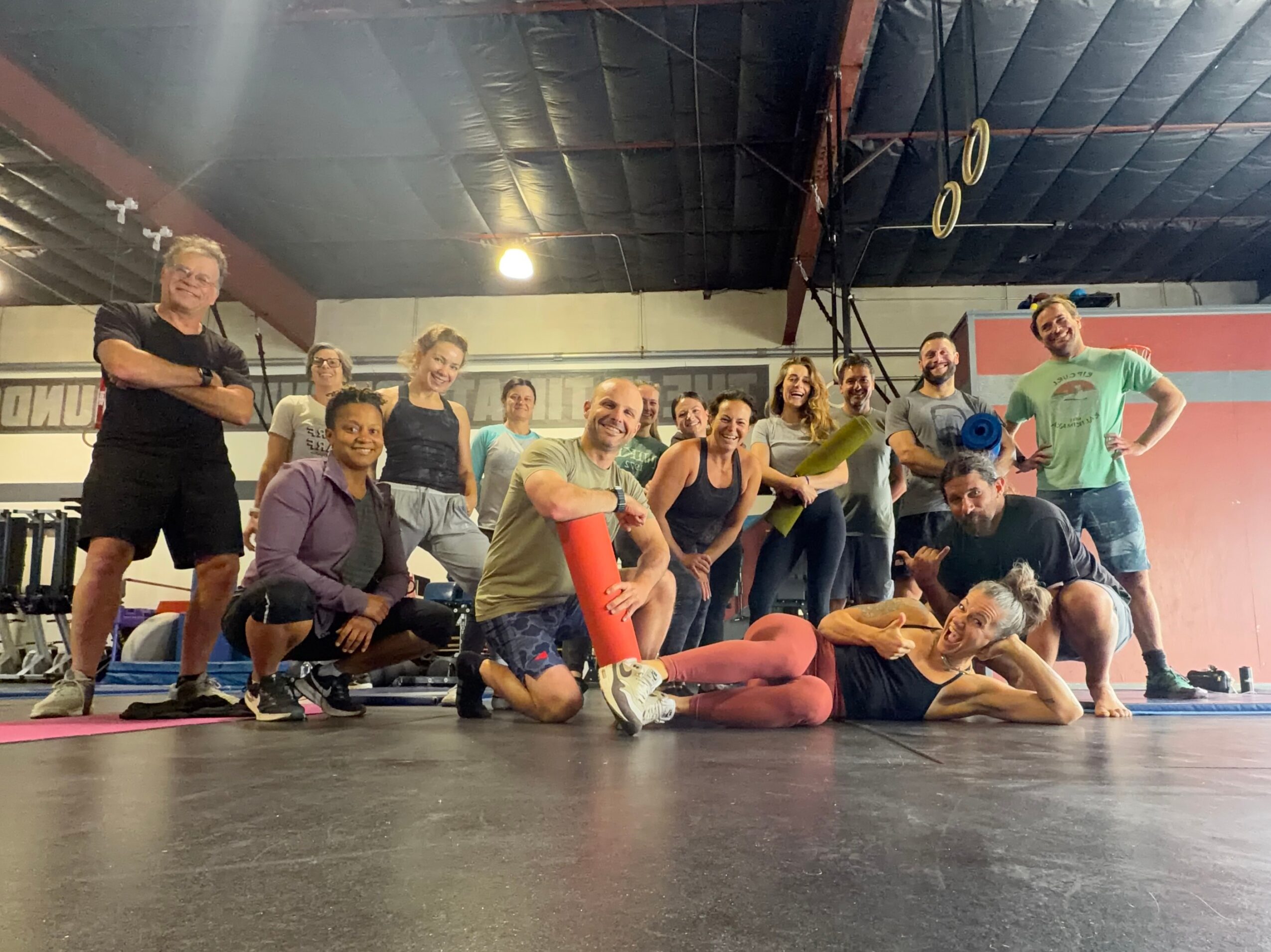 Group of people in a gym, some standing and some sitting or lying on the floor, smiling and posing for a photo.