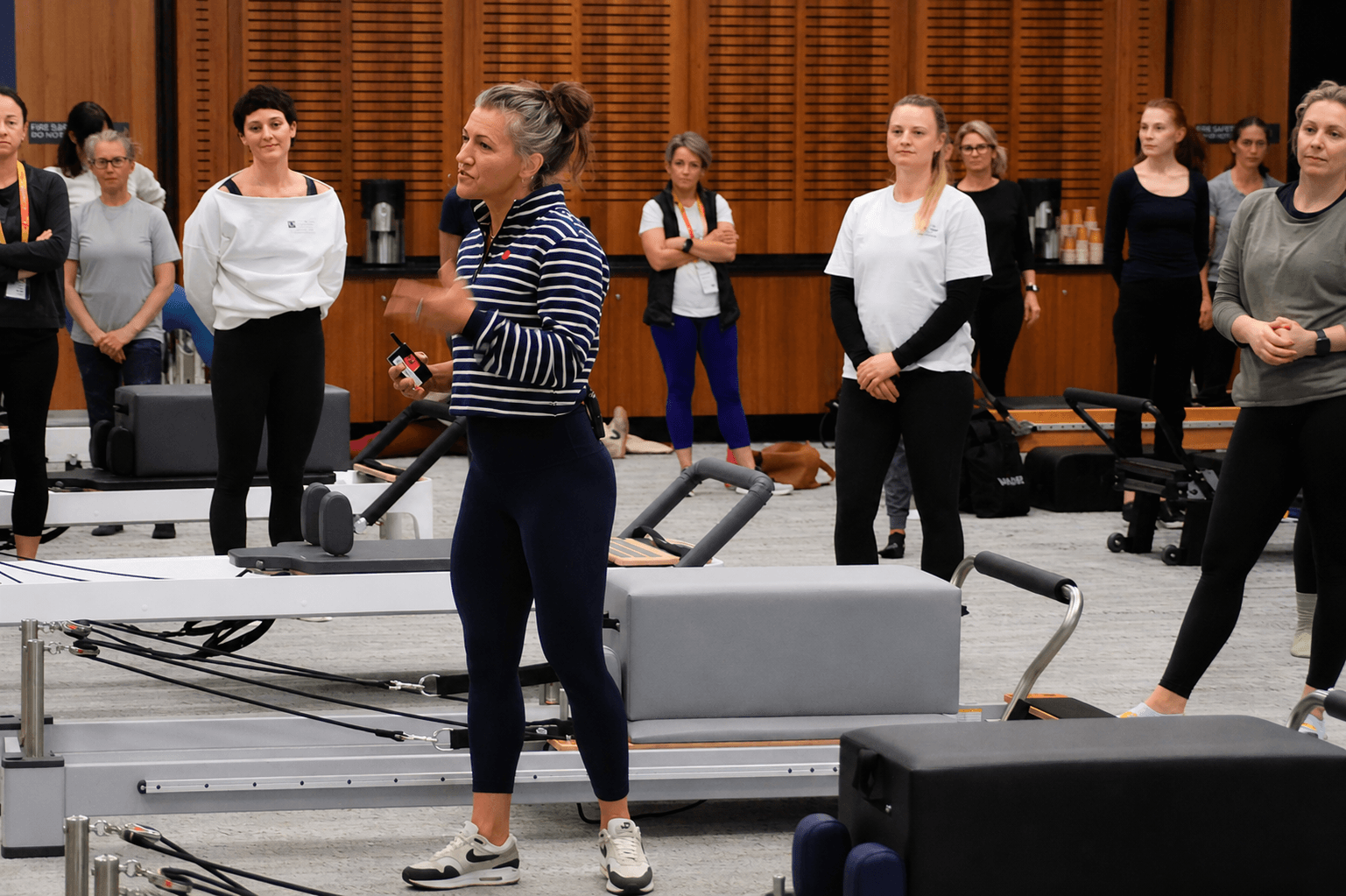 A woman in striped shirt speaking to a group in a gym or fitness studio, with equipment and wooden wall background.