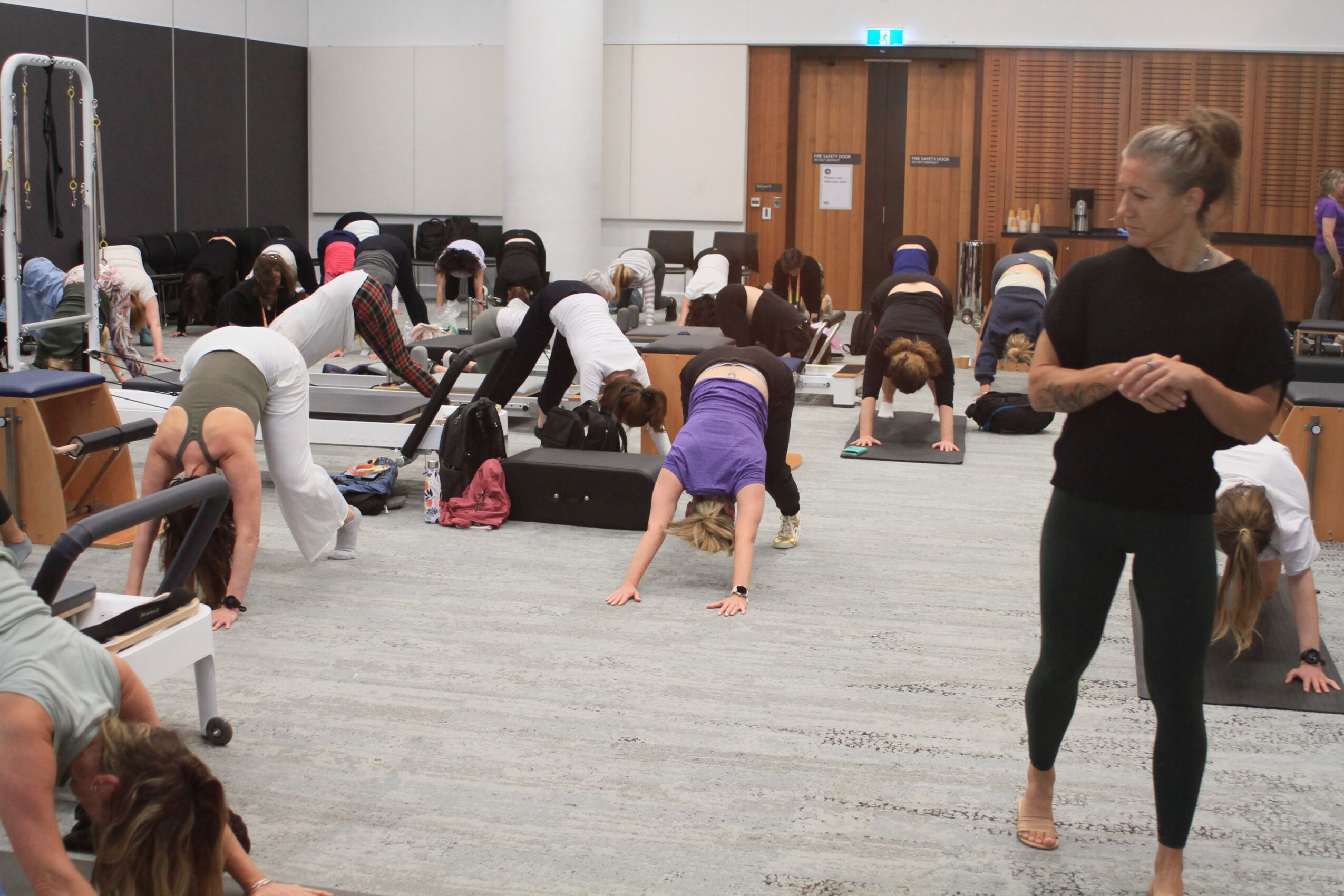 Group of people practicing yoga or stretching in a spacious indoor room with a woman instructor observing, some on mats.