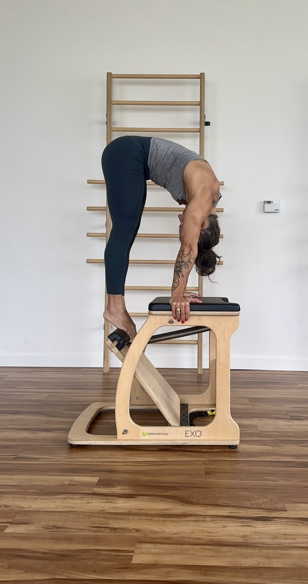Person performing a backbend stretch with hands on a Pilates reformer machine in a room with wooden flooring and wall-mounted exercise equipment.