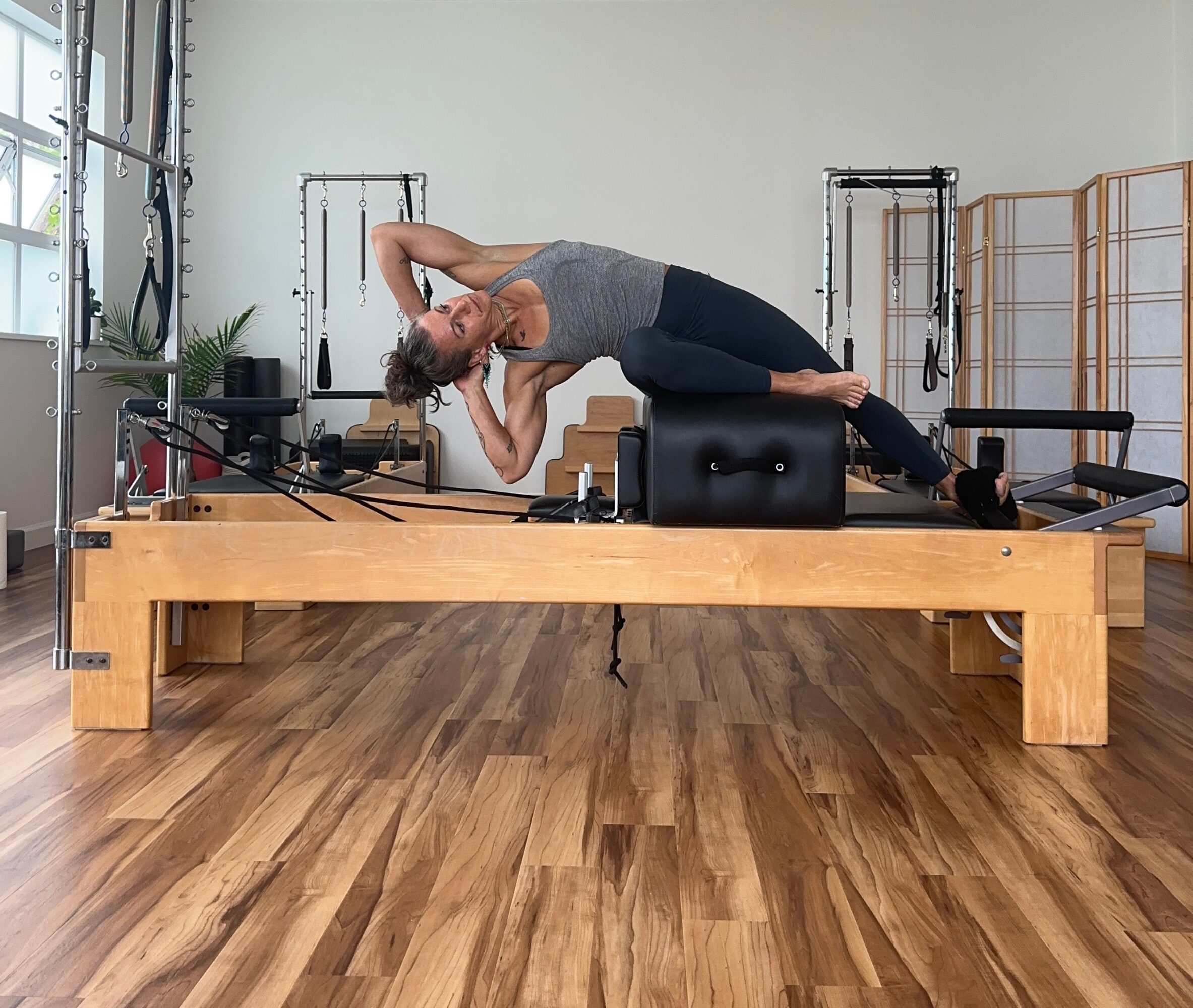 Woman performing side plank exercise on Pilates reformer machine in a fitness studio, with wooden floor and exercise equipment in background.