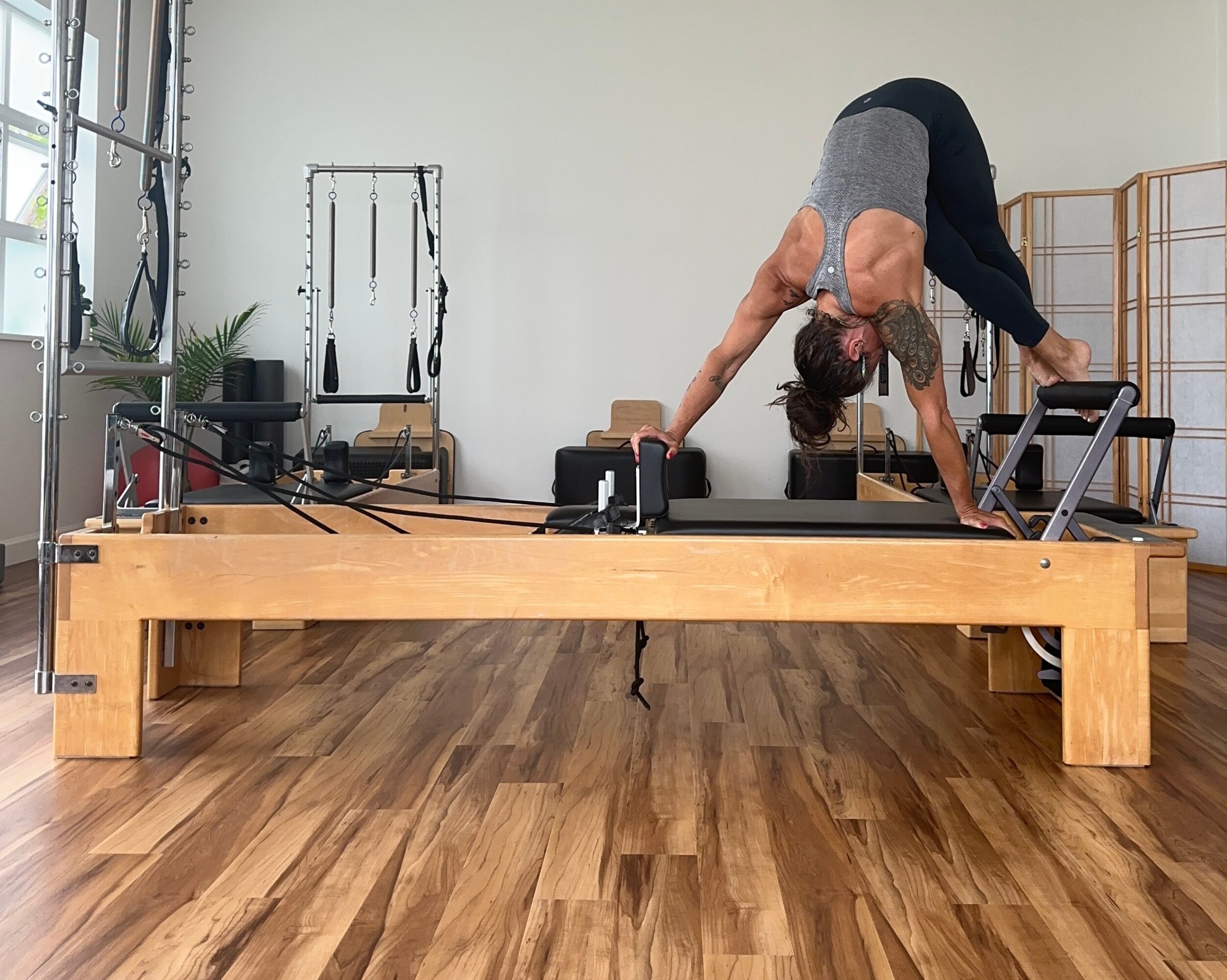 Person performing a stretch on a Pilates reformer machine in a fitness studio, with wooden flooring and exercise equipment in the background.