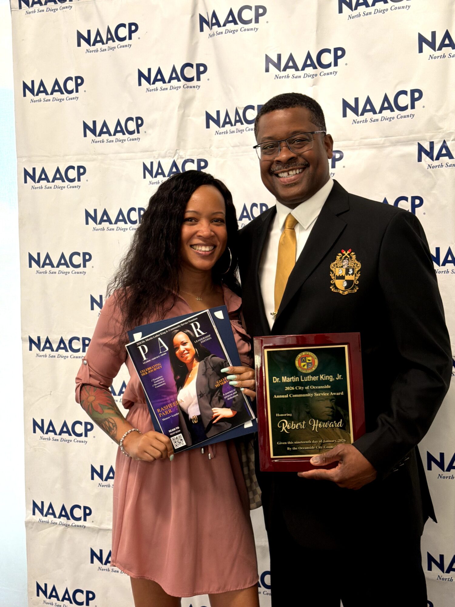 Two people smiling, holding awards, standing in front of a NAACP backdrop, one woman in a pink dress, one man in a suit with a gold tie.