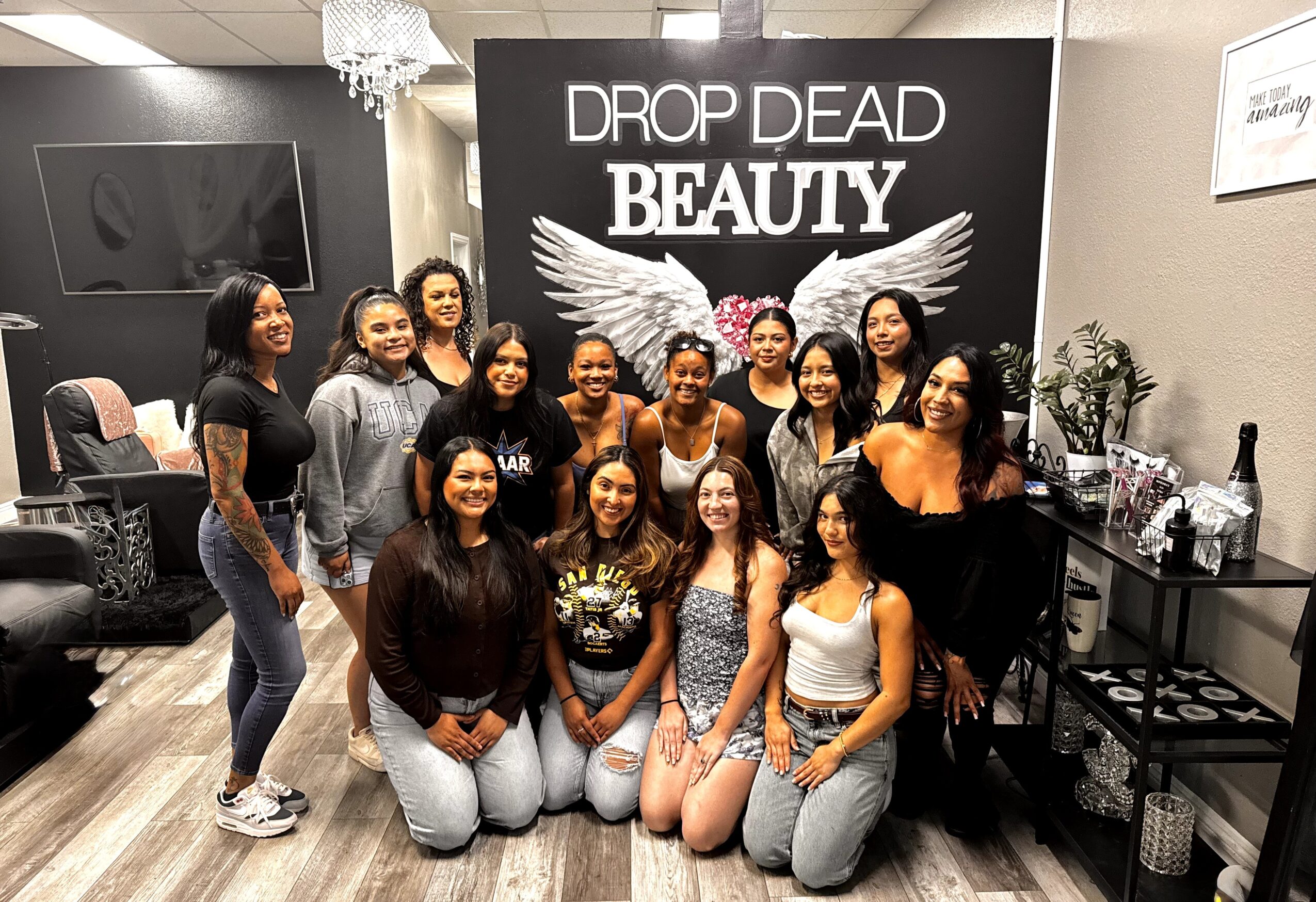 Group of women posing in front of a sign that says 'Drop Dead Beauty' with wings, in a decorated room.