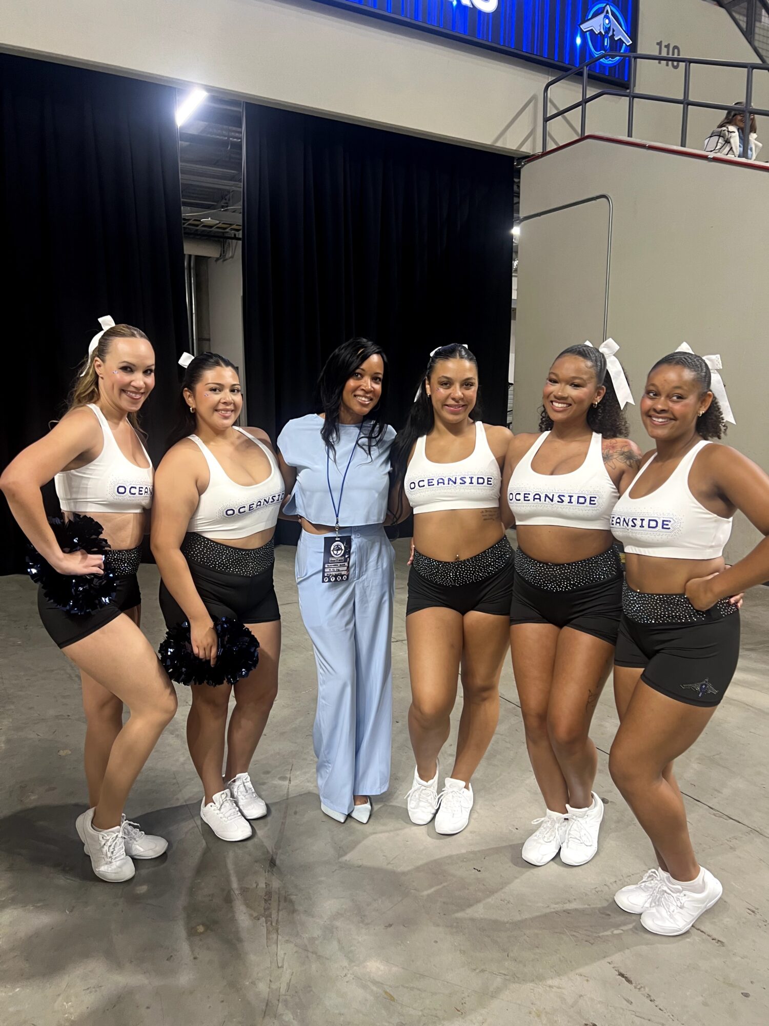 Six women standing together indoors, some in cheerleading outfits, smiling, with a black curtain and a balcony in the background.