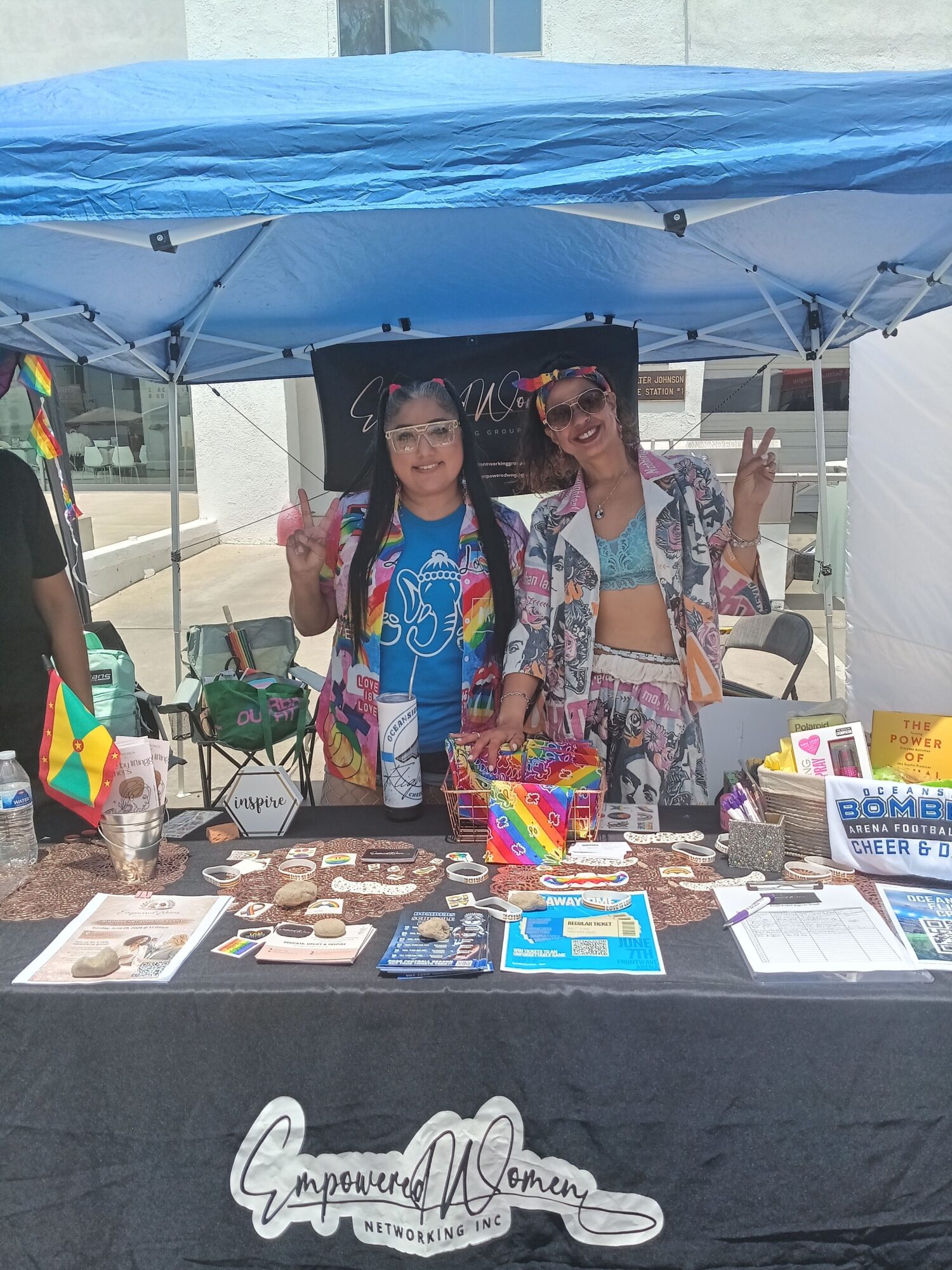 Two women stand behind a table with colorful items and brochures, under a blue canopy at an outdoor event.