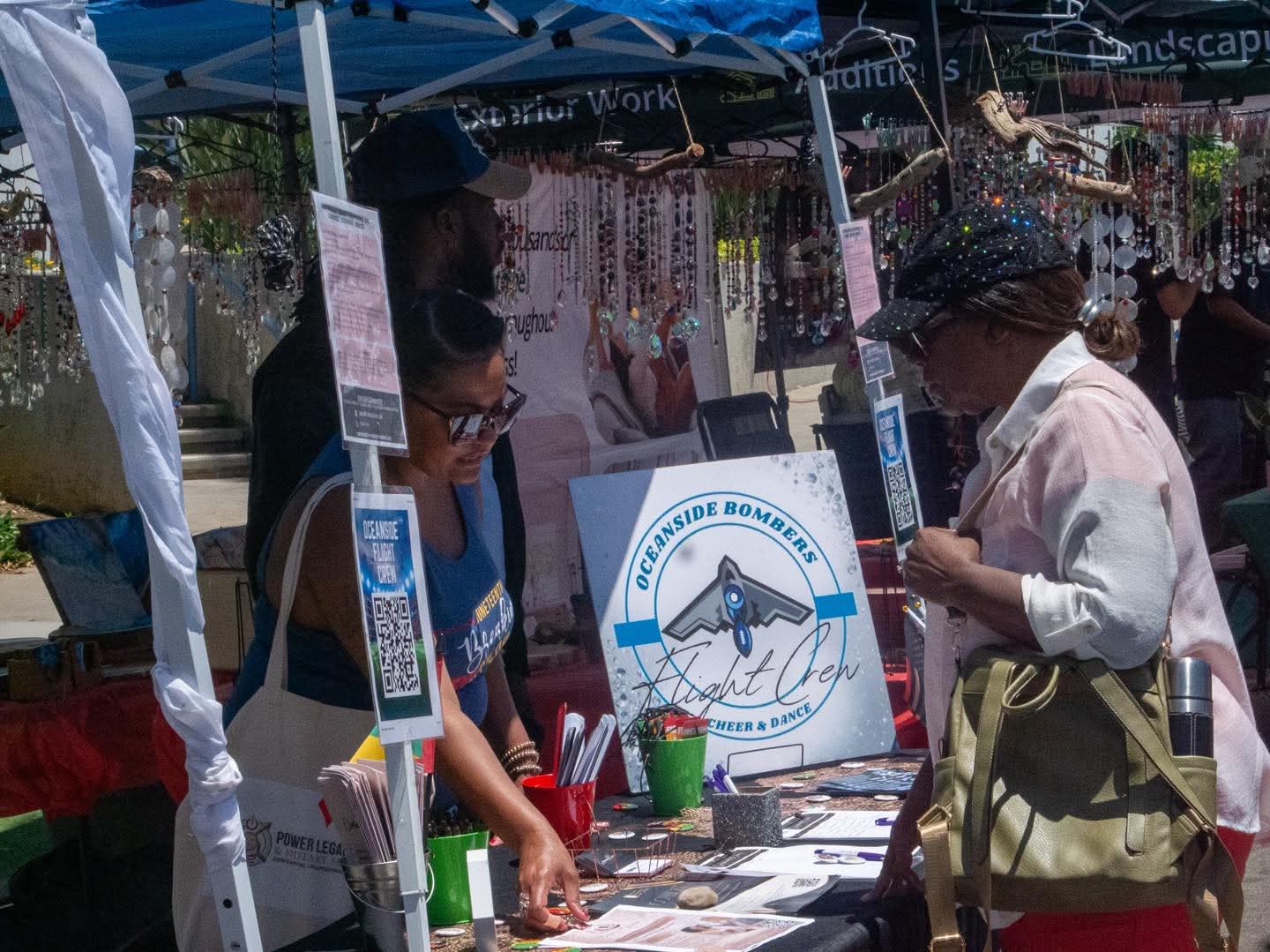 People at an outdoor booth with a sign, browsing items under a canopy, with hanging jewelry displays in background.