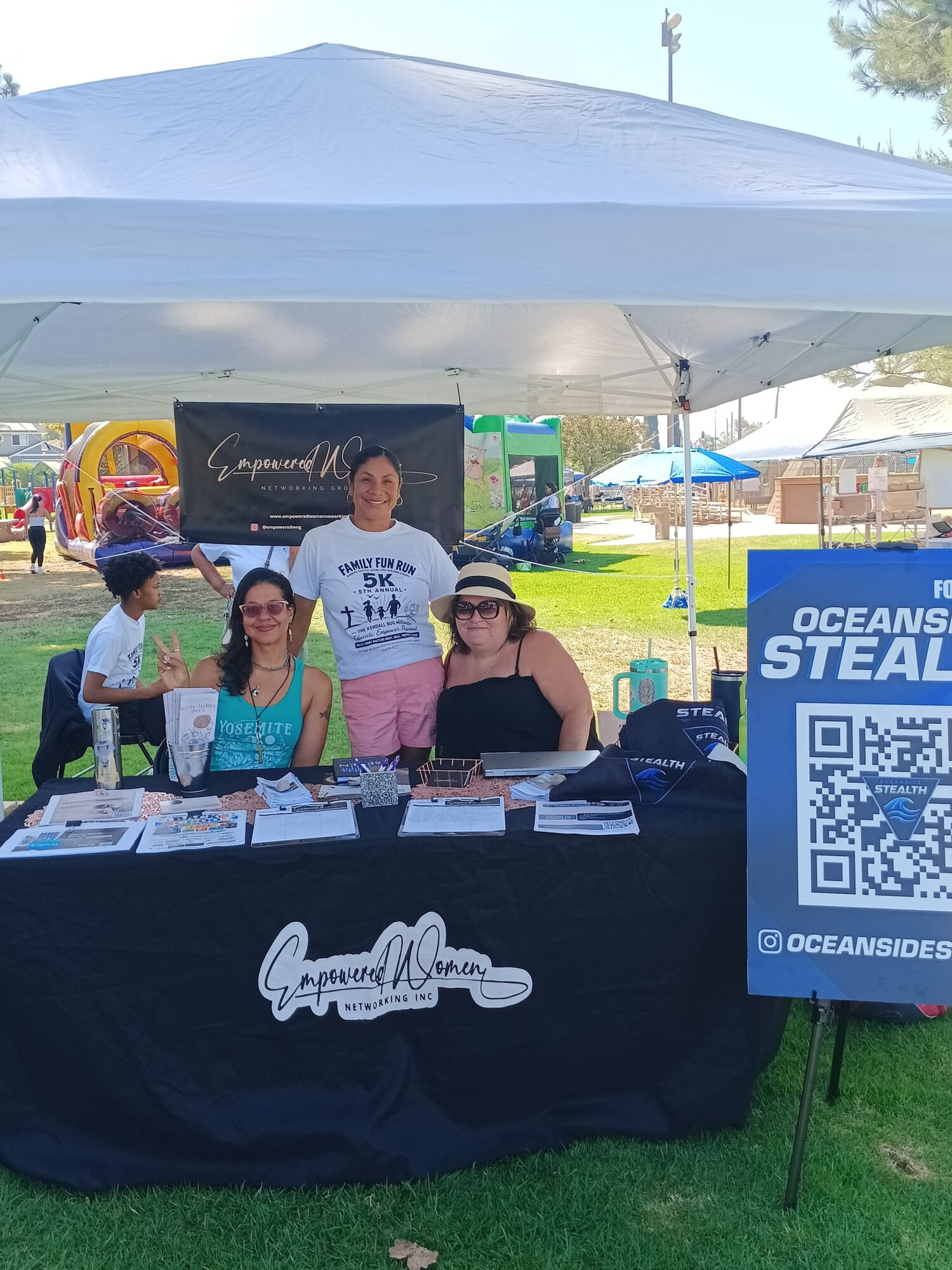 Three people standing behind a table outdoors, with a sign and QR code, under a canopy, on grass, sunny day.
