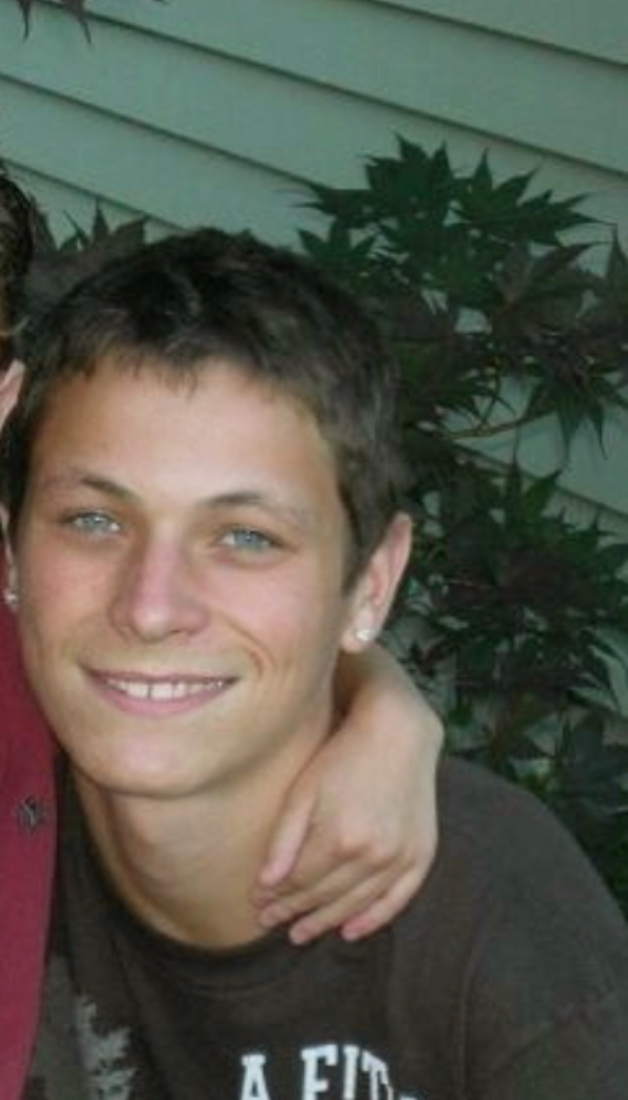 Young man with short dark hair smiling, outdoors with green foliage in background.