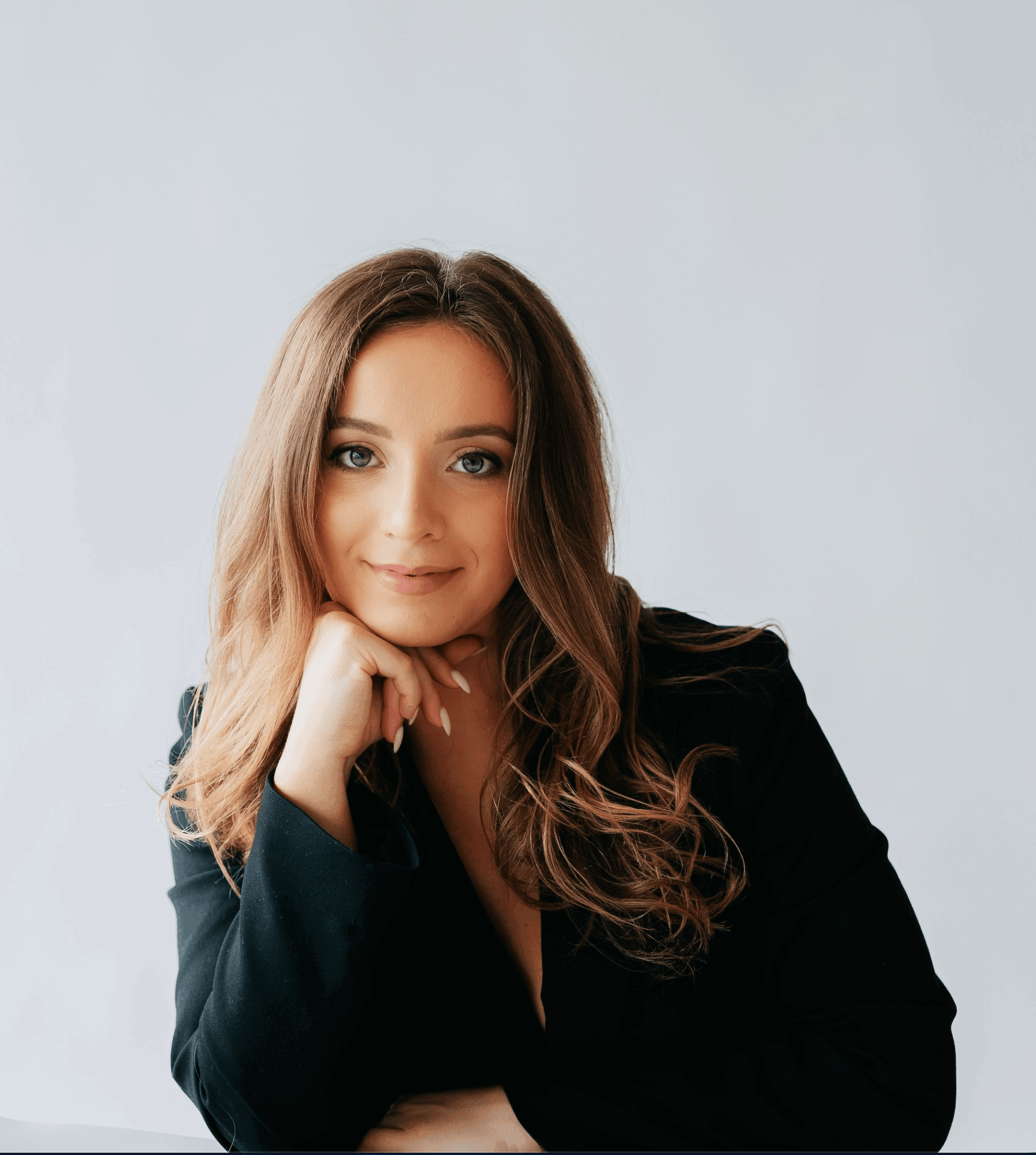 Young woman with long wavy hair, smiling, resting her chin on her hand, wearing a black blazer, against a plain light background.